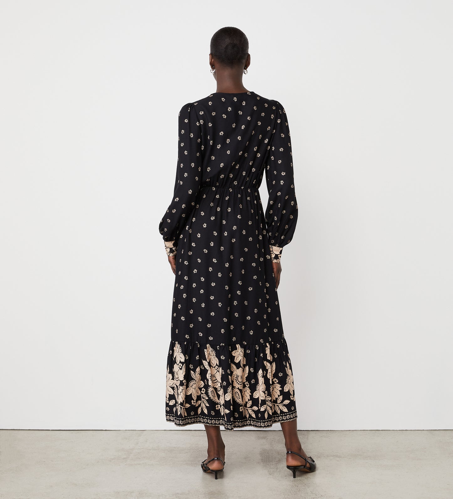 A person stands against a white wall, facing away, wearing the Leyane Black Floral Border Midi Dress with long sleeves, a small floral print, beige hem detailing, and paired with black-heeled sandals.