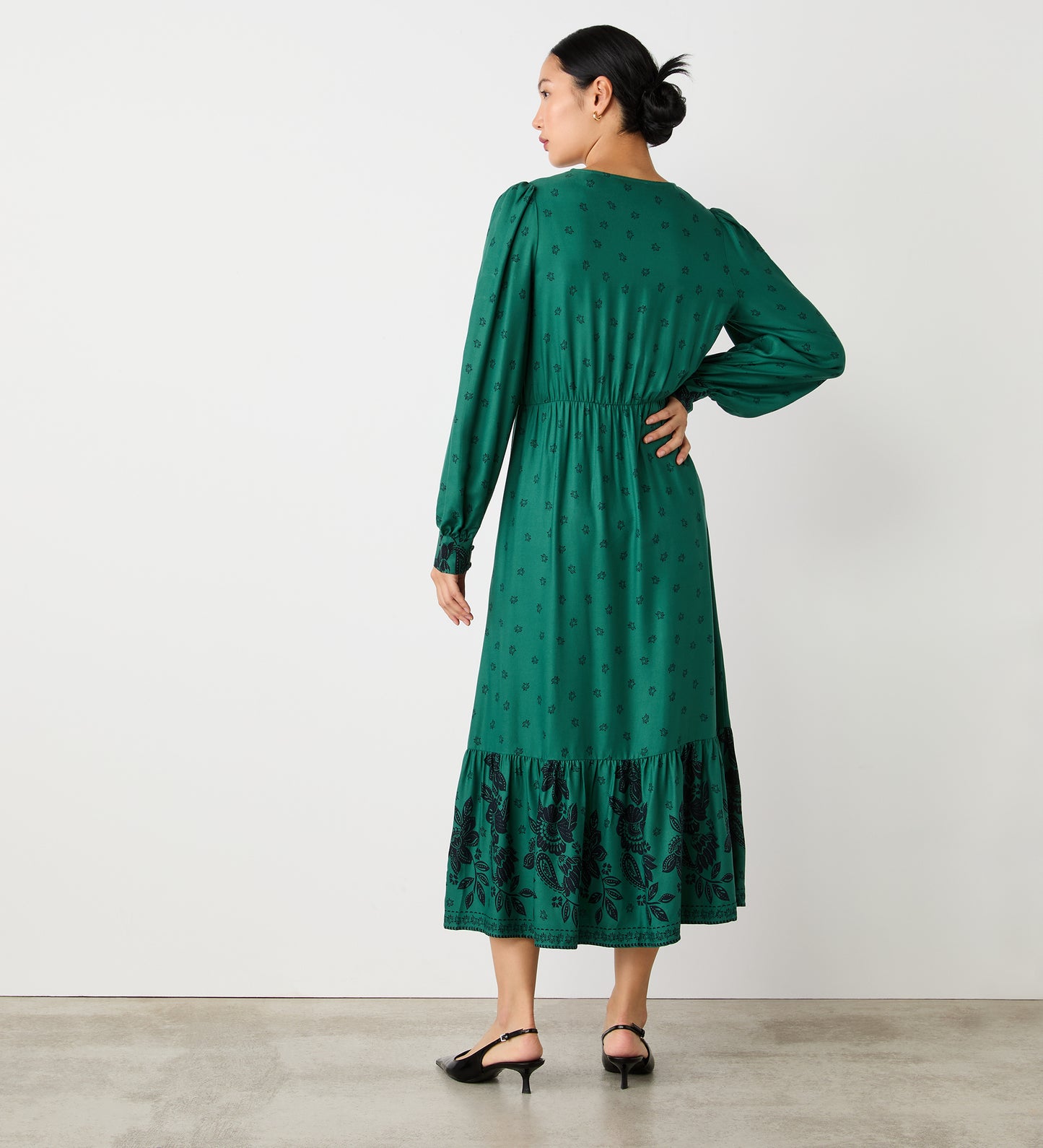 A woman stands indoors facing away, wearing the Leyane Green Floral Border Midi Dress with small patterns and a tiered skirt. She has her hair tied back and pairs the dress with black low-heeled shoes.