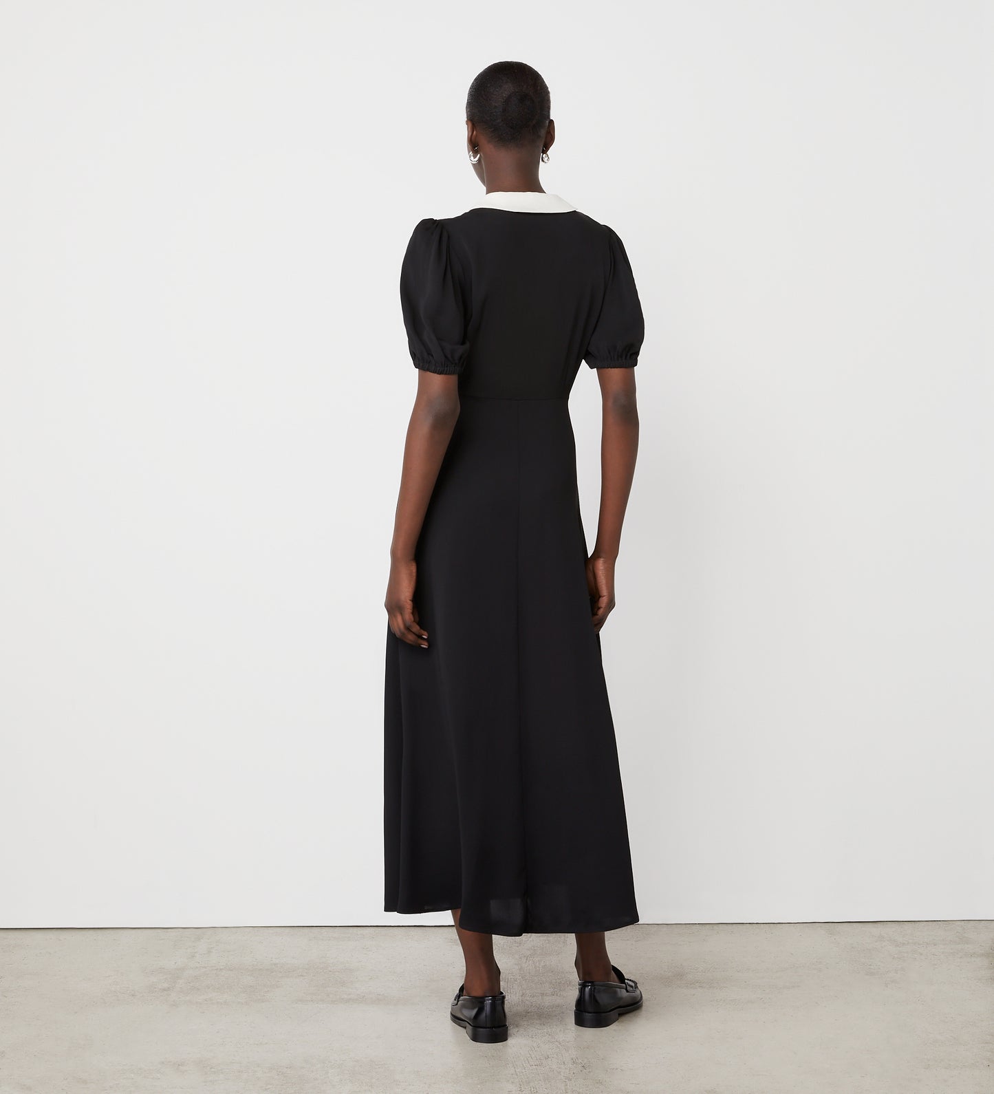 A person with short hair faces a white wall, wearing the Lois Black Crepe Midi Dress with a revere contrast collar, pearl front buttons, and black shoes. The ankle-length dress is shown from behind in a minimalist setting.