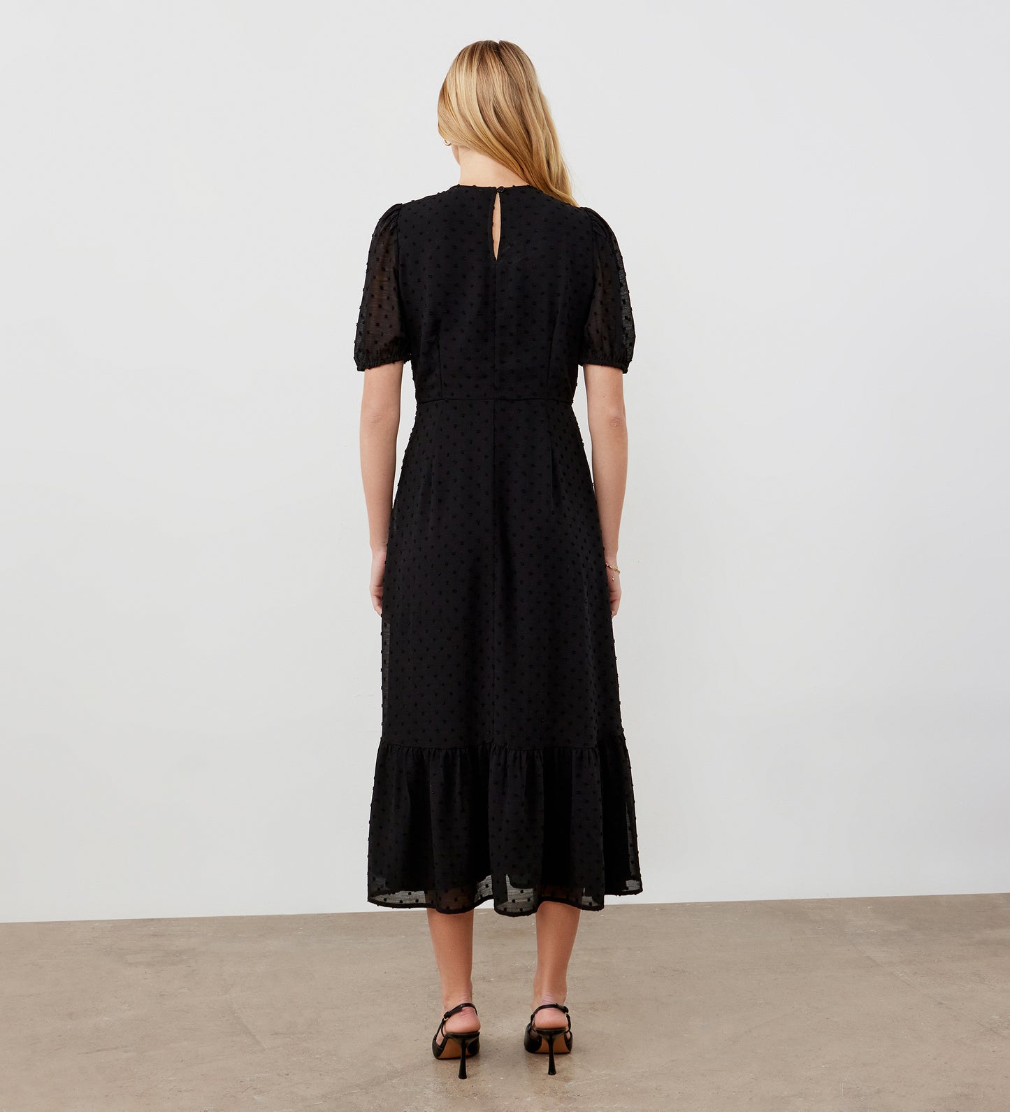 A woman with long blonde hair faces away, wearing the Loretta Black Georgette Midi Dress featuring a V-neck, textured dot pattern, ruffled hem, and black heeled sandals, set against a plain light background.