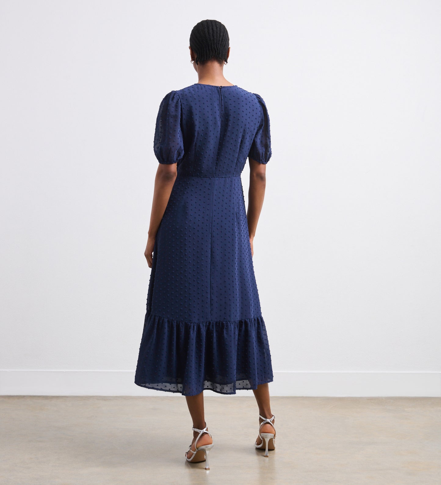 A person stands indoors facing away, wearing the Loretta Navy Georgette Midi Dress featuring a V-neck, puffed short sleeves, fitted waist, and flared tiered hemline. They have short hair and strappy heeled sandals.