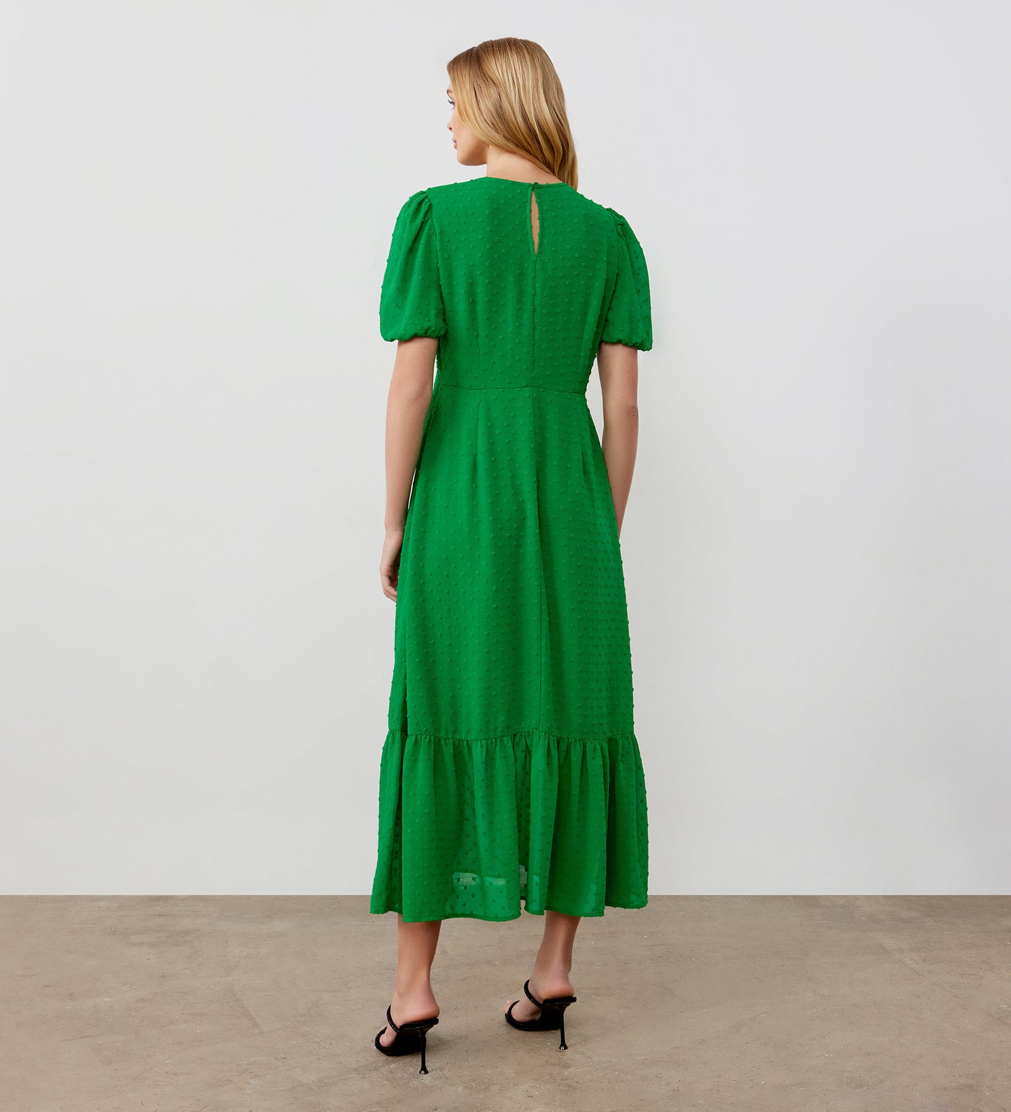 A woman with blonde hair, facing away against a plain light background, wears the Loretta Green Georgette Midi Dress featuring a ruffled hem and black heeled sandals.