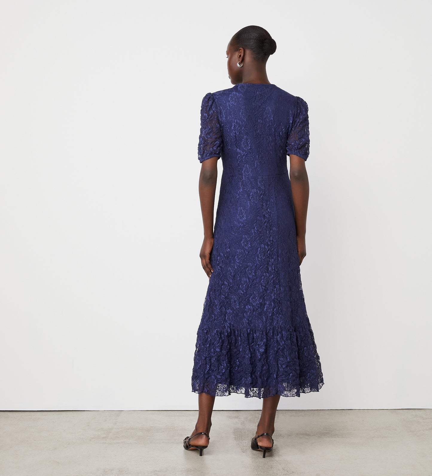 A woman faces away in the Loretta Navy Lace Midi Dress, featuring an empire waist and ruffled hem. Her hair is in a low bun, and she wears black heeled sandals against a plain white wall.