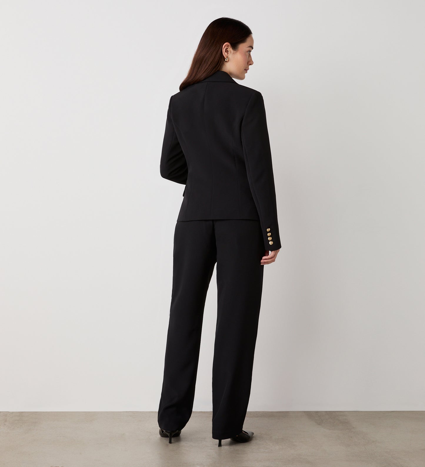 A woman stands with her back to the camera, wearing the Lydia Black Blazer featuring gold button details on the sleeves and black heels, set against a plain white wall and concrete floor.