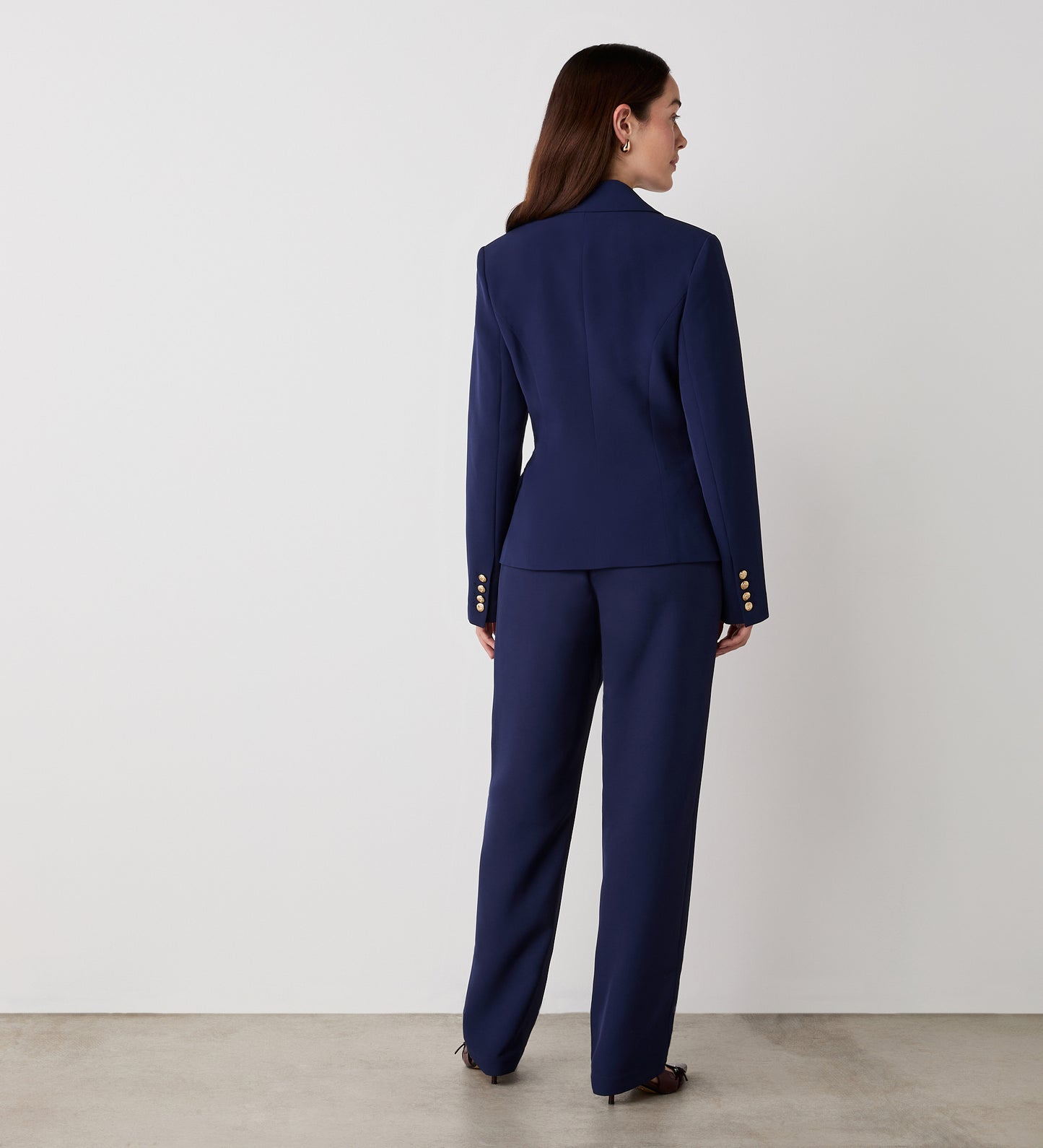 A woman stands facing away in the Lydia Navy Blazer with gold buttons and matching pants. Her long brown hair flows down her back as she poses against a plain white wall on a concrete floor.