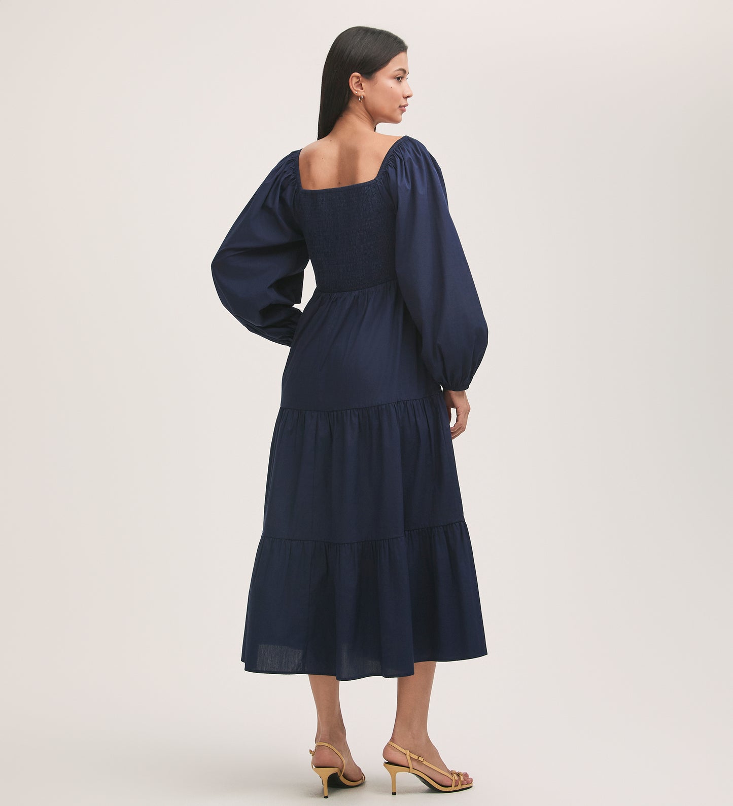 A woman with long dark hair faces away, wearing the Lauretta Navy Cotton Midi Tiered Dress with a square neckline and puff sleeves. The fit-and-flare design pairs with beige heeled sandals against a plain, light background.