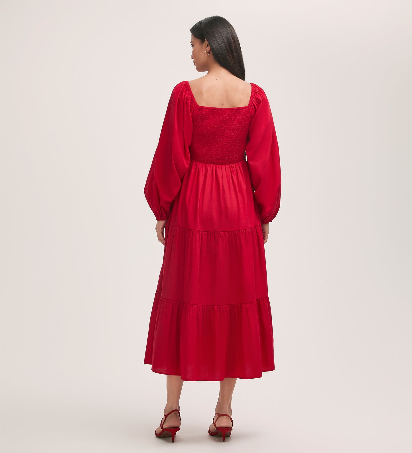 A woman stands with her back to the camera, wearing the Lauretta Red Cotton Midi Tiered Dress with puffed sleeves and a shirred bodice, paired with red heeled sandals against a plain light background.