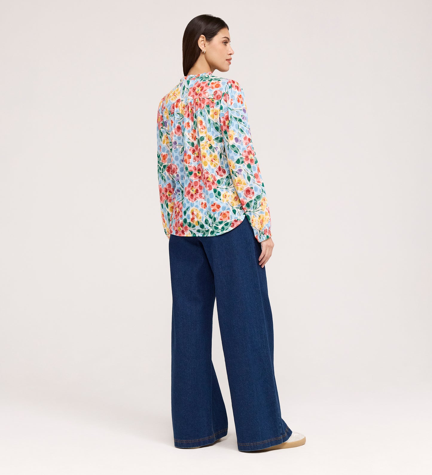 A woman with straight, dark hair wears the Lizzie Blue Floral Jersey Blouse, featuring long sleeves and a tie neck, paired with wide-leg blue jeans and beige shoes. She stands facing away against a plain light background.