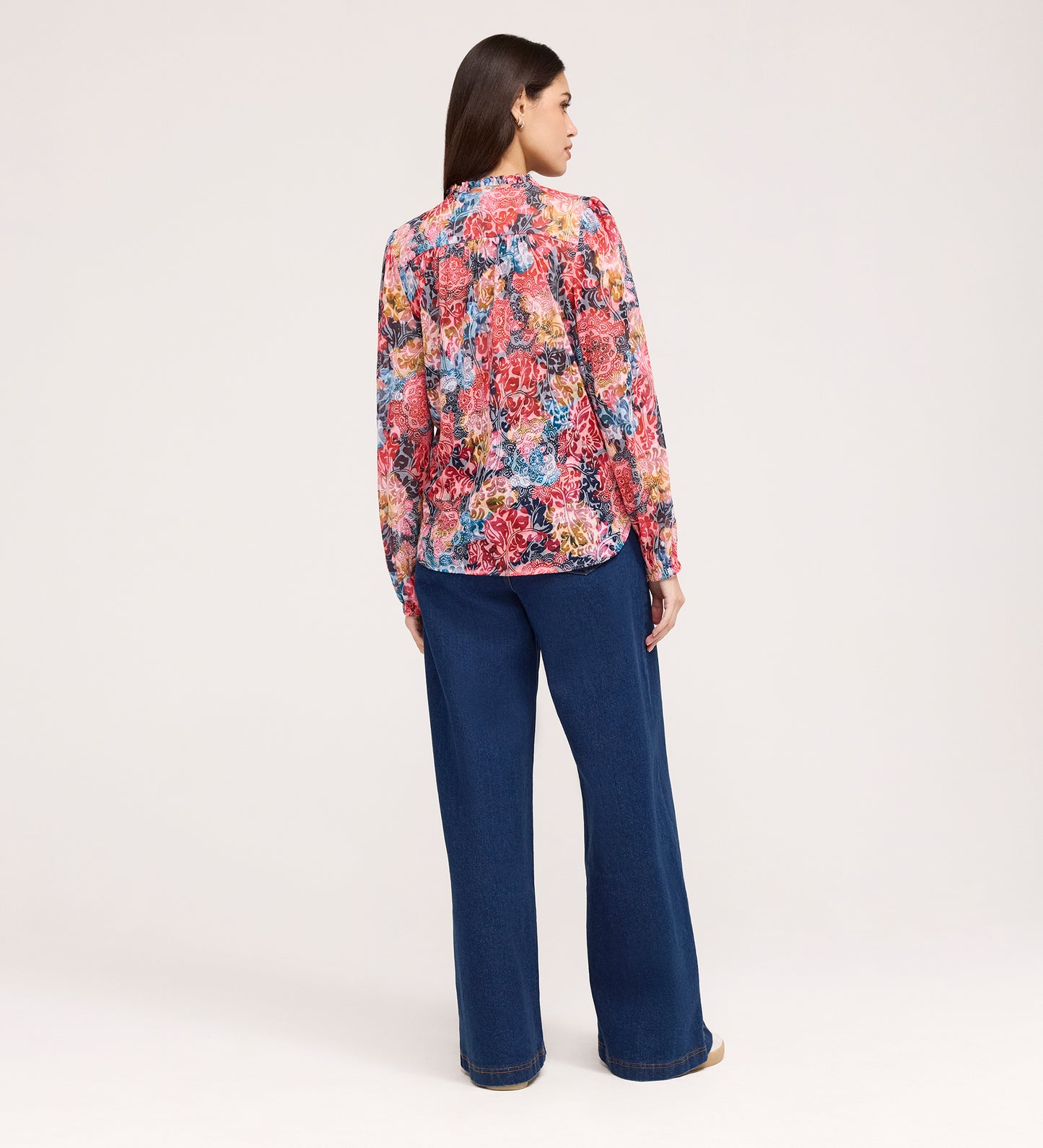 A woman stands facing away, wearing the Lizzie Pink Floral Jersey Blouse with blouson sleeves and wide-leg blue jeans against a plain light background.