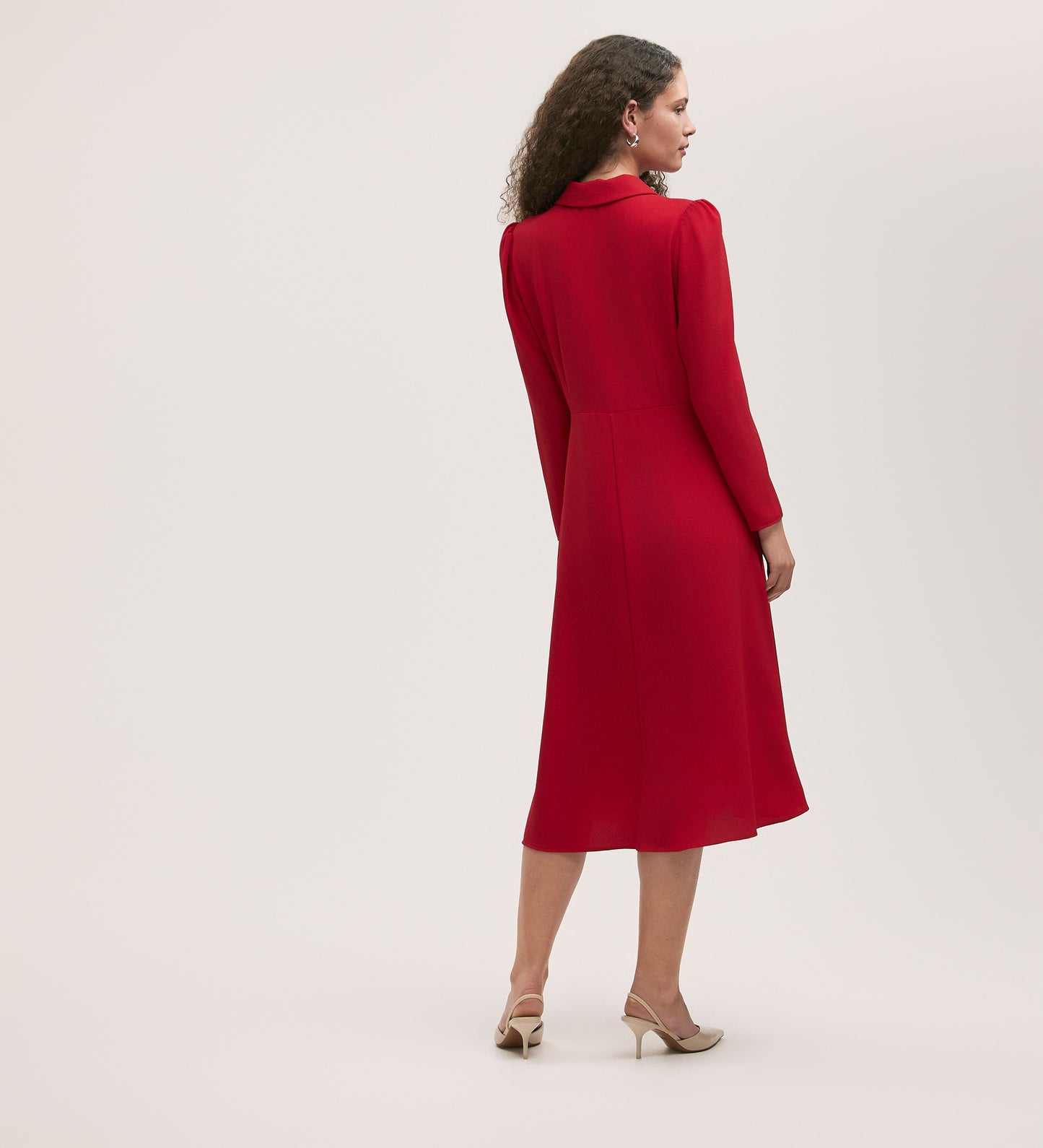 A woman with curly hair models the Lucia Red Crepe Midi Dress, featuring long sleeves and a fitted waist, shown from the back. She pairs it with beige heels and stands against a plain light background for an elegant look.