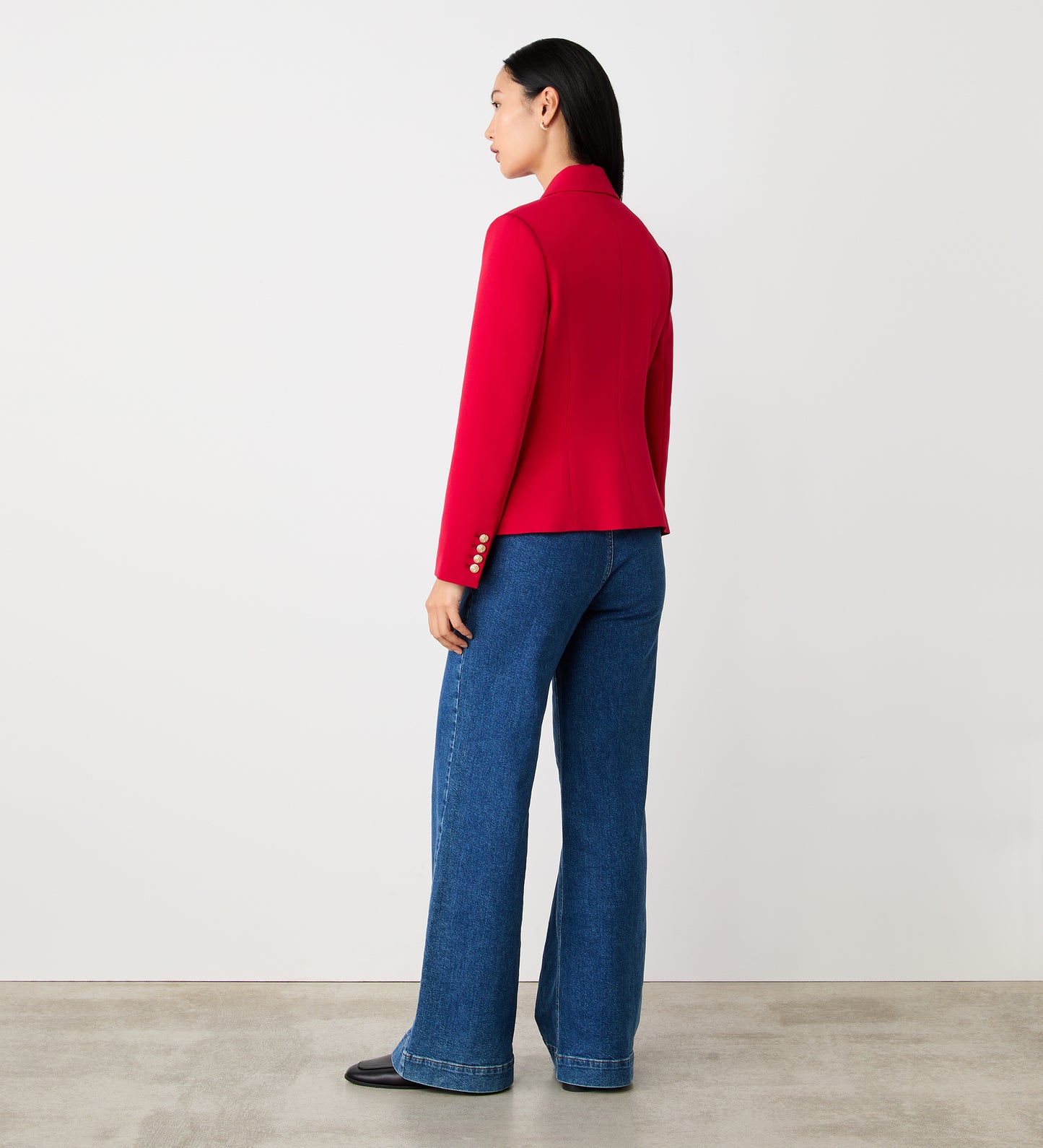 A woman faces away from the camera, wearing the Lydia Red Blazer with gold buttons, paired with blue wide-leg jeans and black shoes. She poses against a plain white wall on a concrete floor.