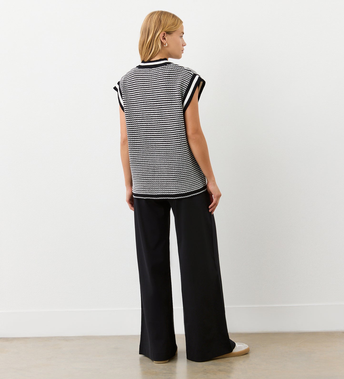 A person with shoulder-length blonde hair, seen from behind, wears the Maree Black Sleeveless Cardigan and loose black pants, standing against a plain white wall and light floor.