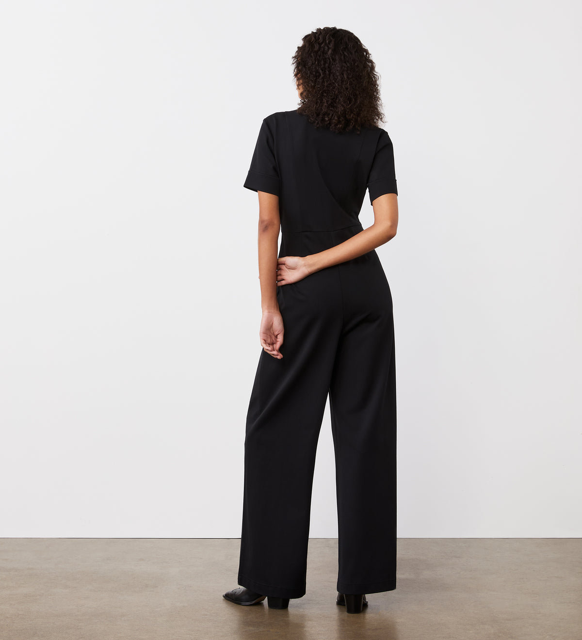 A person with curly hair stands against a white wall, facing away, wearing the Marley Black Ponte Jersey Jumpsuit with short sleeves and black shoes. One arm is bent behind their back.