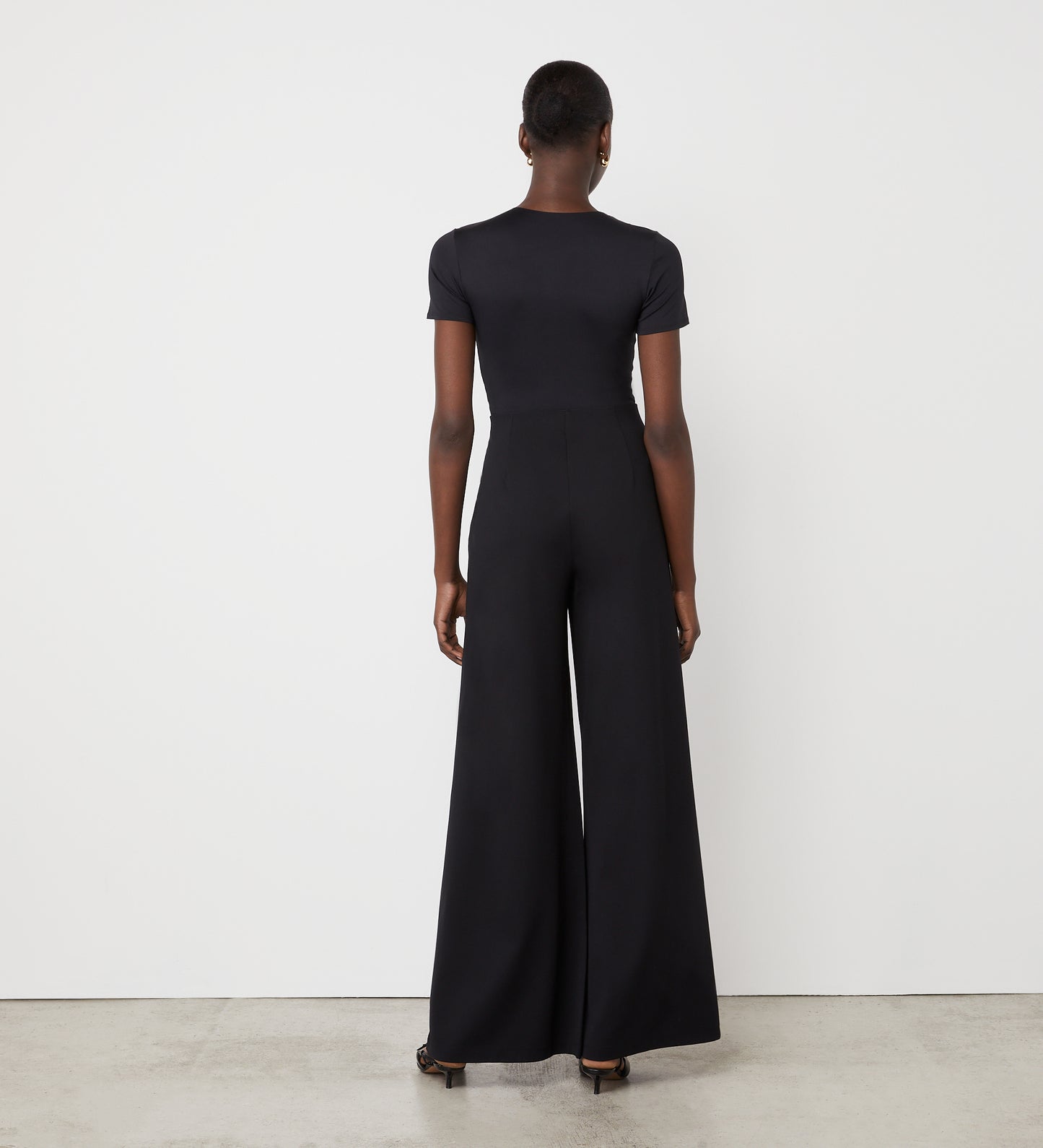 A person stands facing a white wall, wearing a fitted black top and the Matilda Black Ponte Jersey Wide Leg Trousers with an elasticated waistband, paired with black heels. The photo is shot from behind in a minimalist setting.