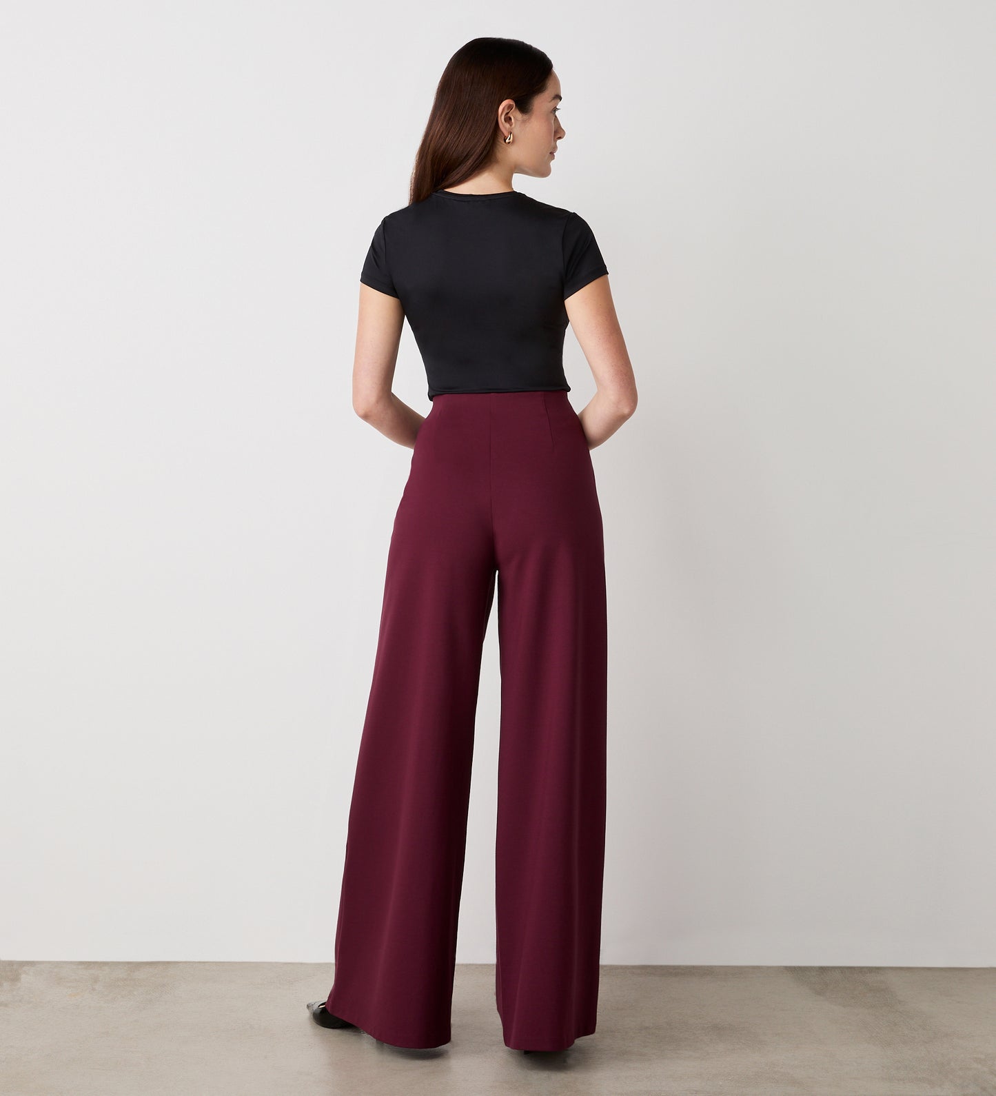 A woman faces away from the camera, wearing a fitted black short-sleeve top and the Matilda Burgundy Ponte Jersey Wide Leg Trousers with an elasticated waistband, in a minimalist indoor setting with a plain white wall and concrete floor.