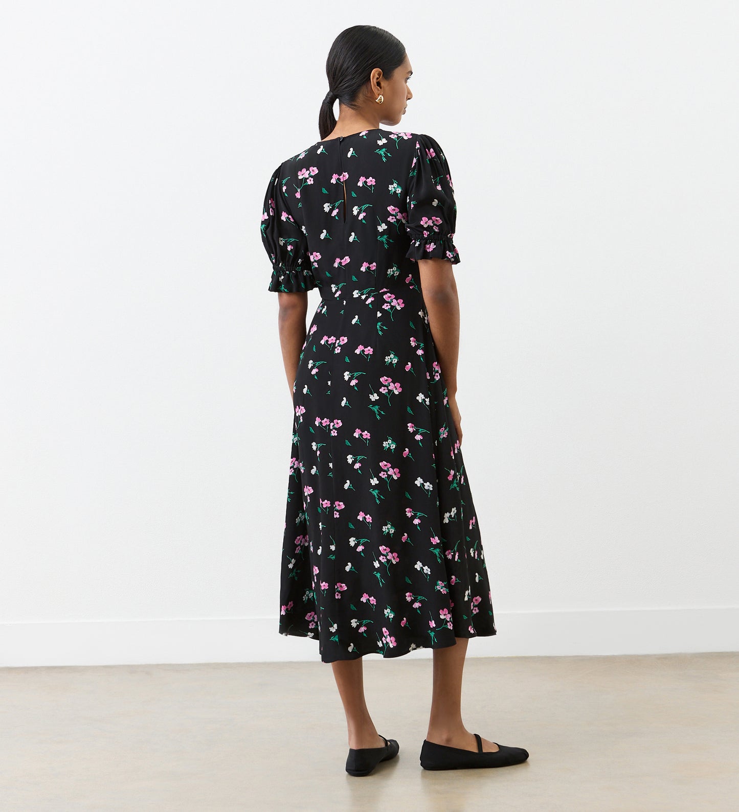 A woman stands indoors with her back turned, wearing the Mela Black Floral Crepe Midi Dress featuring pink and white floral patterns, puffed short sleeves, and an empire waist. Her hair is pulled back as she faces a white wall on a light floor.