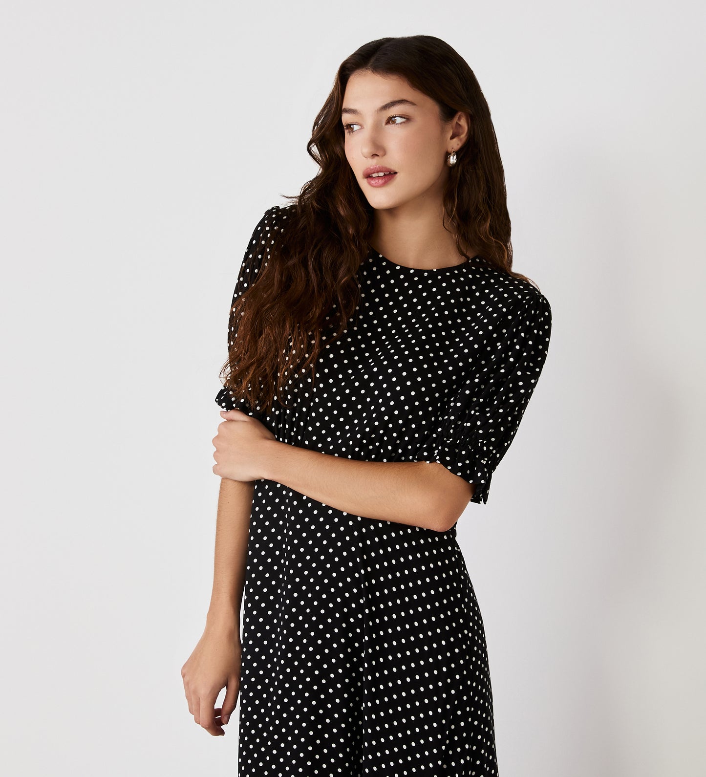 A woman with long wavy brown hair wears the Mela Black Polka Dot Crepe Midi Dress, featuring an empire waist, as she stands against a plain white background and gazes to the side with a neutral expression.