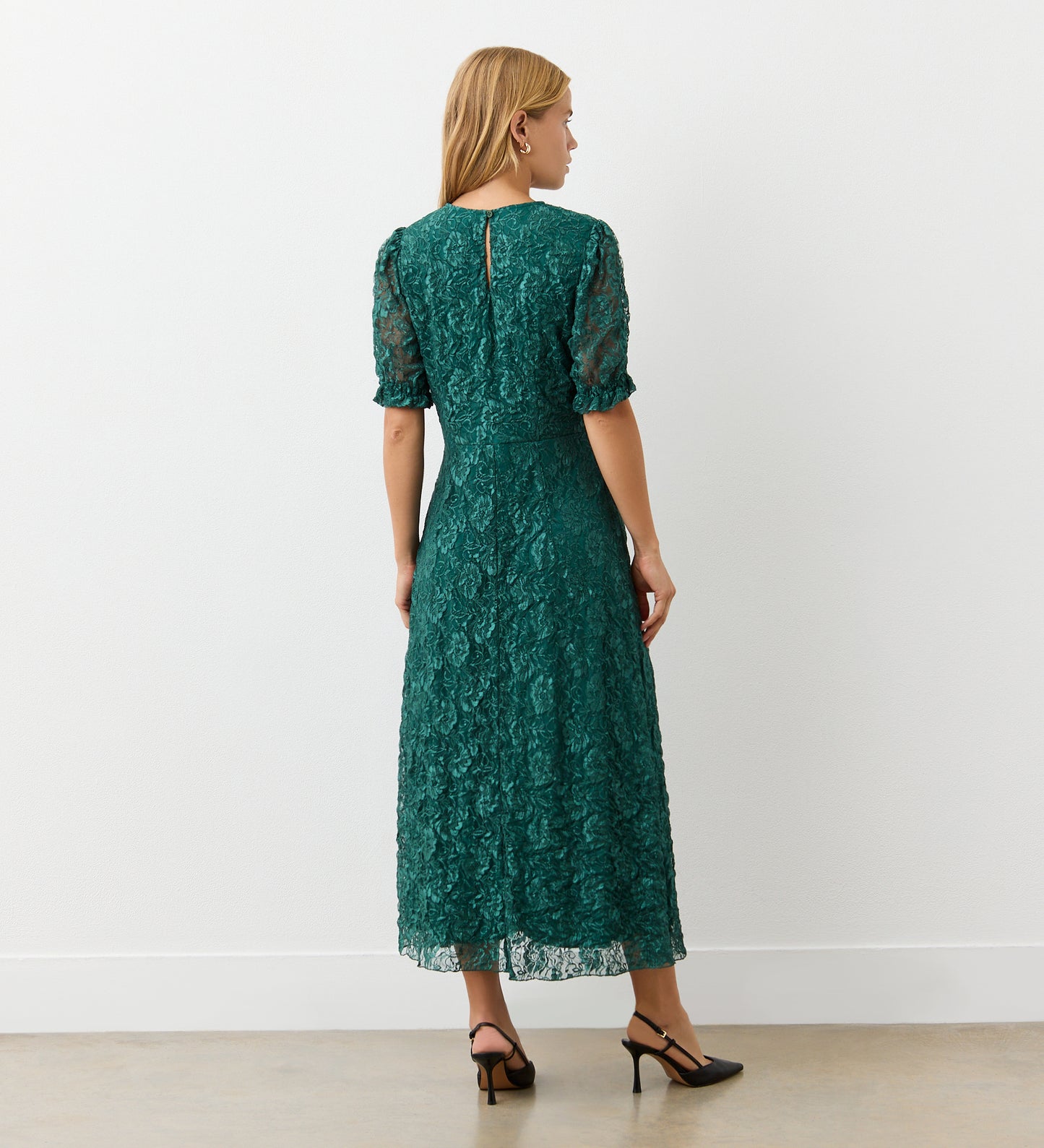 A woman with long blonde hair faces away, wearing the Mela Green Lace Midi Dress with an elegant neckline and short sleeves, paired with black heels. She stands against a plain white wall on a light-colored floor.