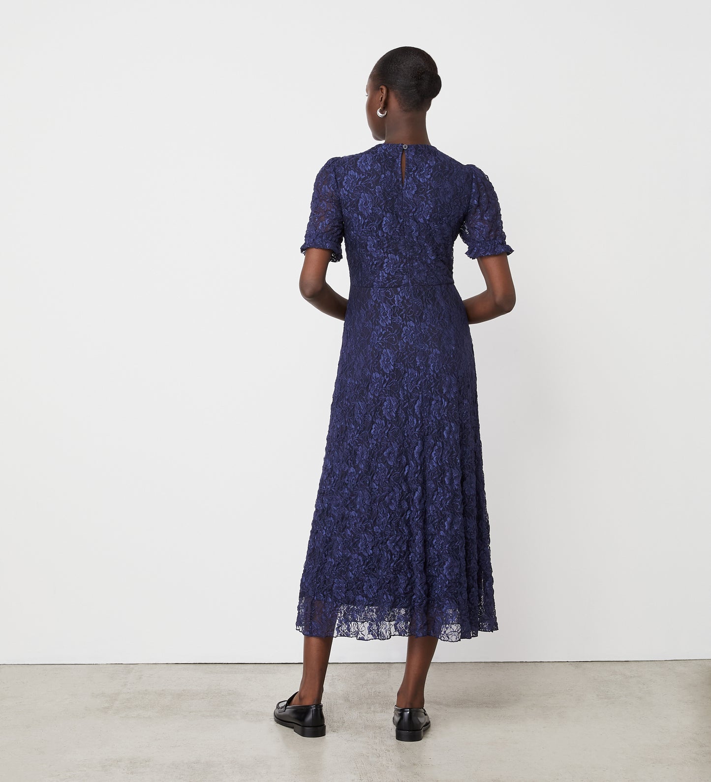 A person faces away wearing the Mela Navy Lace Midi Dress, featuring a lace fabric, elegant round neckline, and short sleeves. They pair it with black shoes against a plain white wall and light polished floor.
