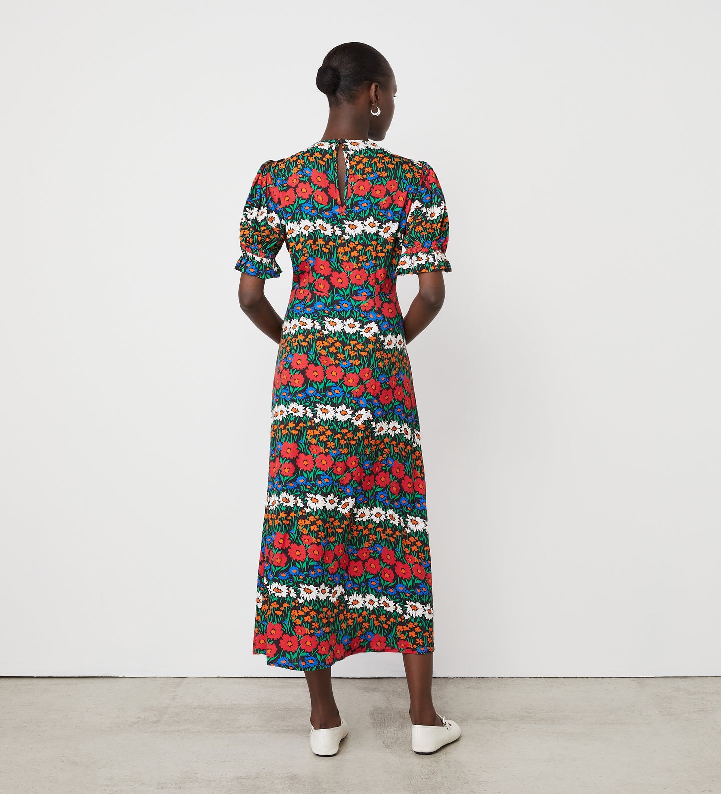 A woman faces away, wearing the Mela Red Floral Stripe Crepe Midi Tea Dress with short puff sleeves and an empire waist. Her hair is in a bun, paired with white shoes against a plain white wall and concrete floor.