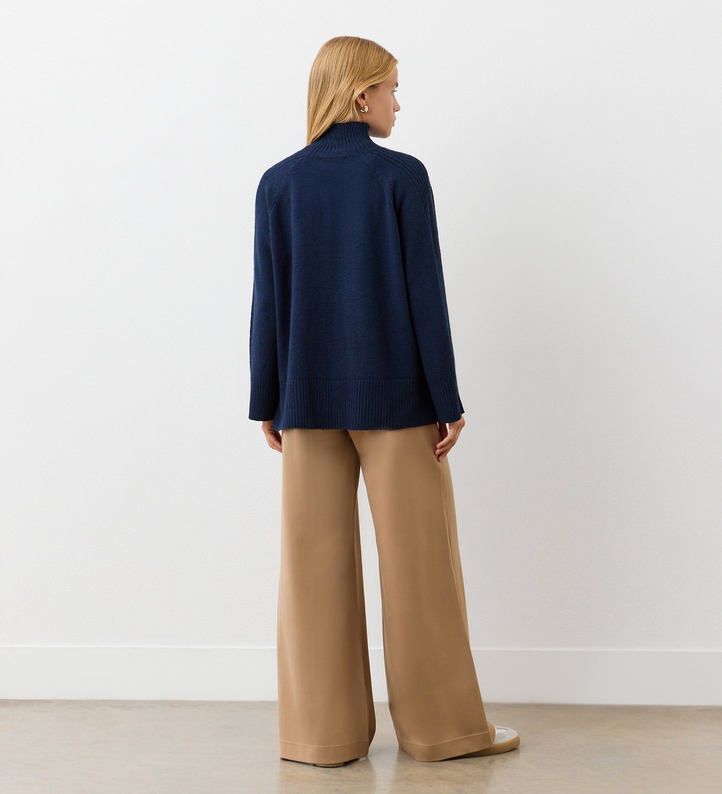 A person with long blonde hair faces away, wearing the Melanie Navy Knitted Jumper with ribbed detailing and wide-legged beige pants, paired with beige shoes against a plain white wall and light-colored floor.