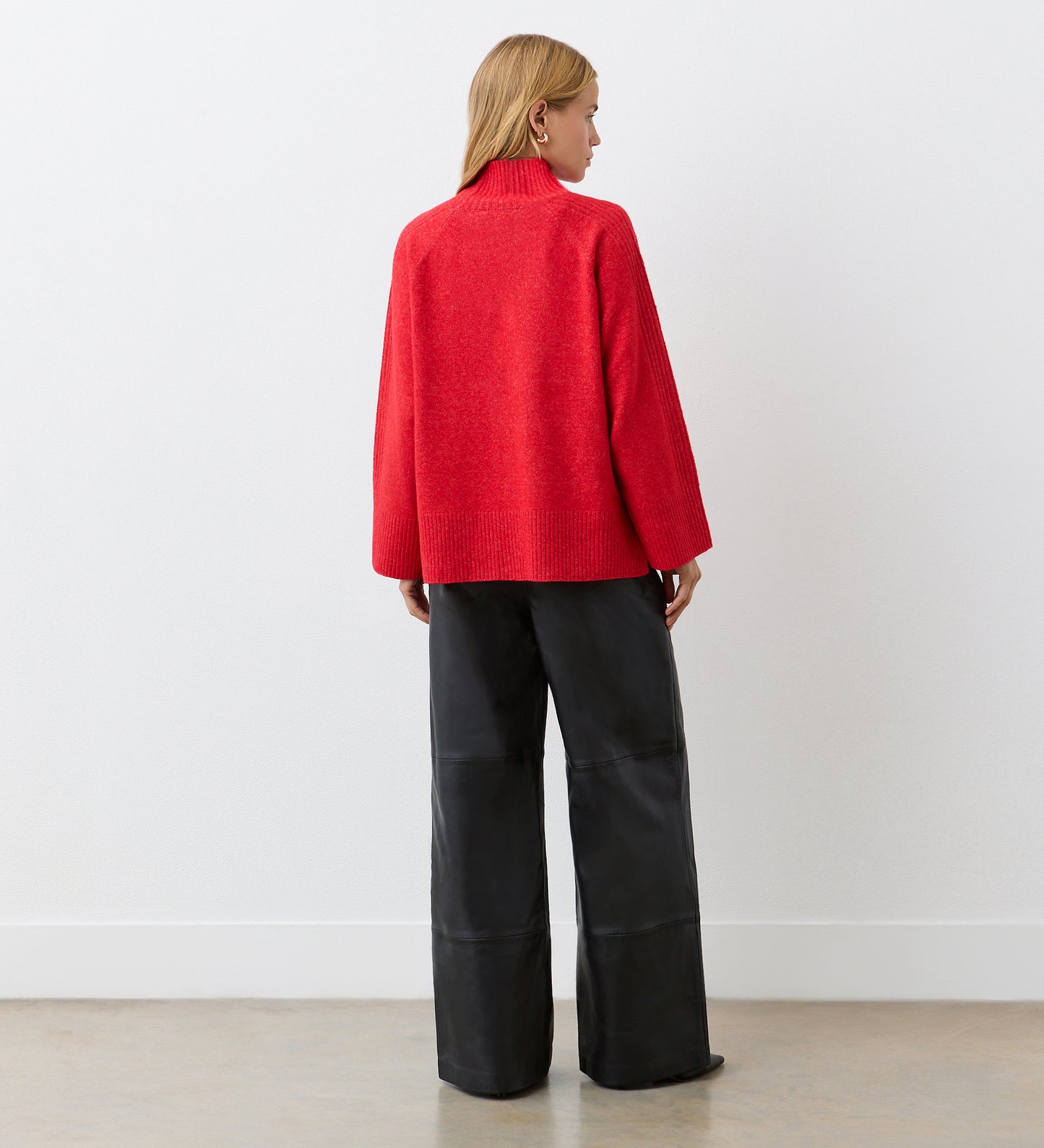 A woman with straight, light brown hair faces away, wearing the Melanie Red Knitted Jumper—an oversized chunky knit with ribbed detailing—paired with wide-leg black pants. She stands against a plain white wall on a light floor.