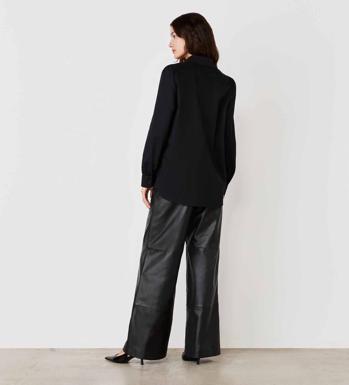 A woman stands facing away, wearing the Mimi Black Cotton Rich Shirt and wide-leg leather pants. She is on a light floor against a plain white wall, her head turned slightly to the side.