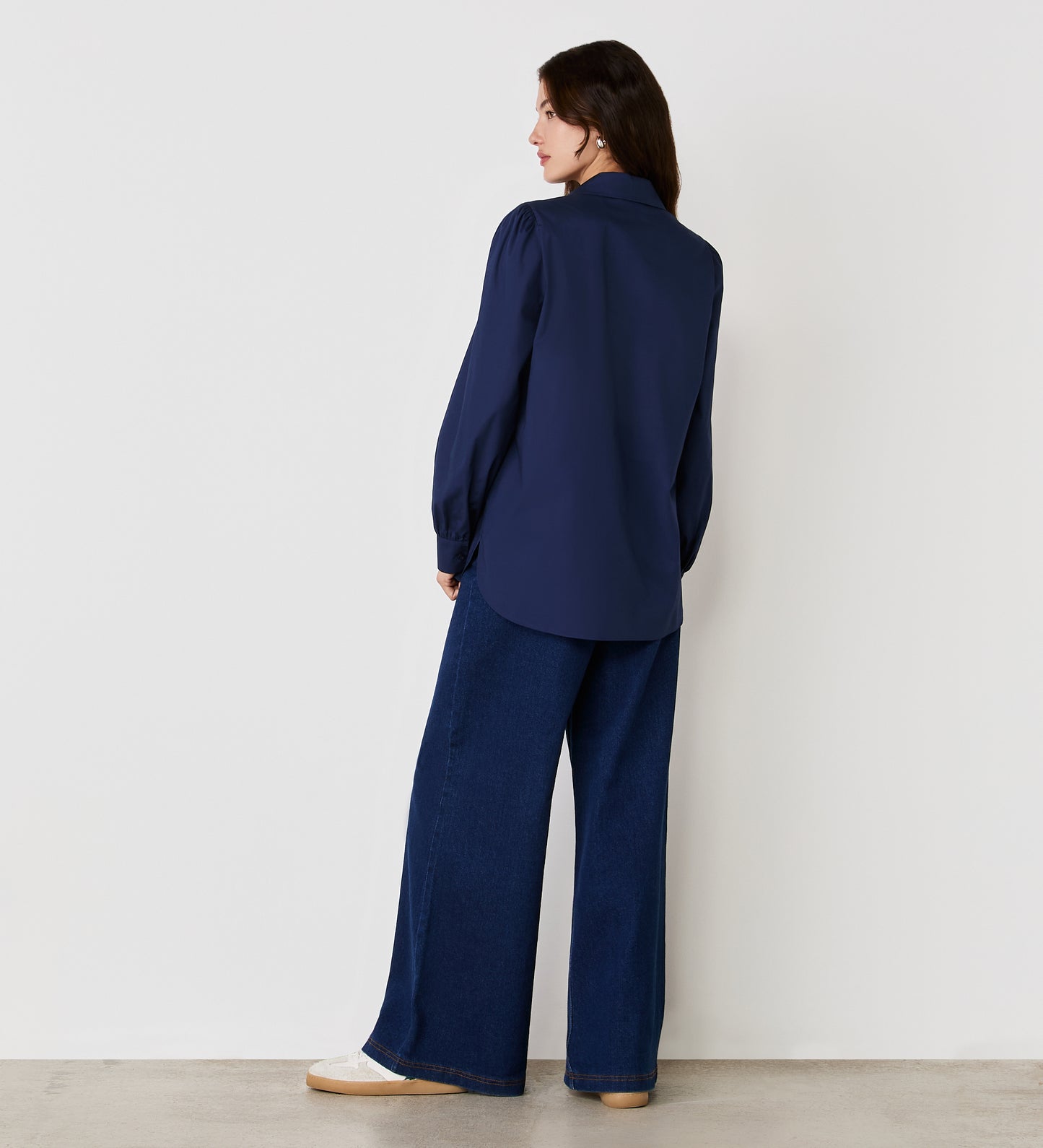 A woman stands facing away from the camera against a white wall, wearing the Mimi Navy Cotton Rich Shirt and wide-leg blue jeans.