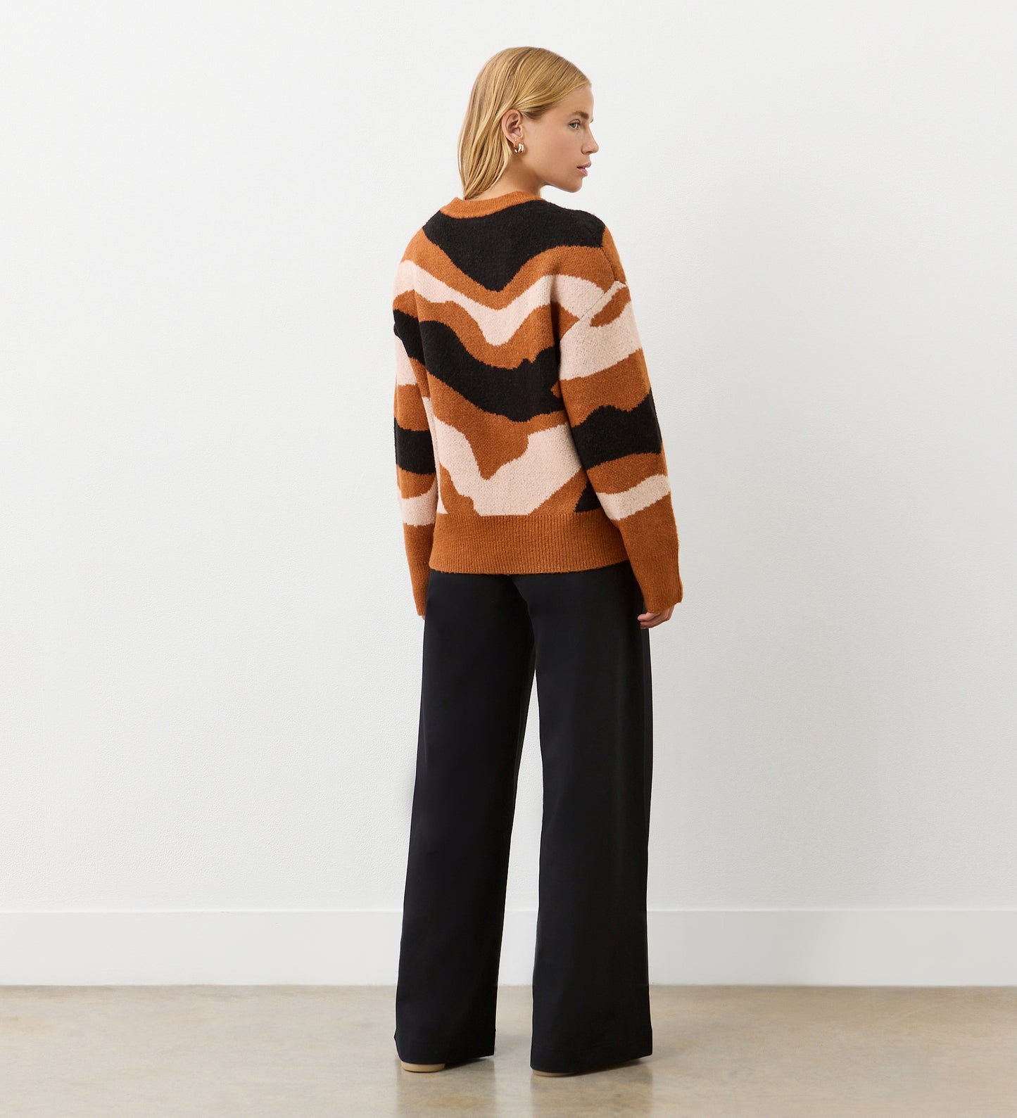 A woman with short blond hair faces away, wearing the Minnie Brown Abstract Print Knitted Jumper and loose black trousers. The abstract print stands out against the plain white wall and light-colored floor.