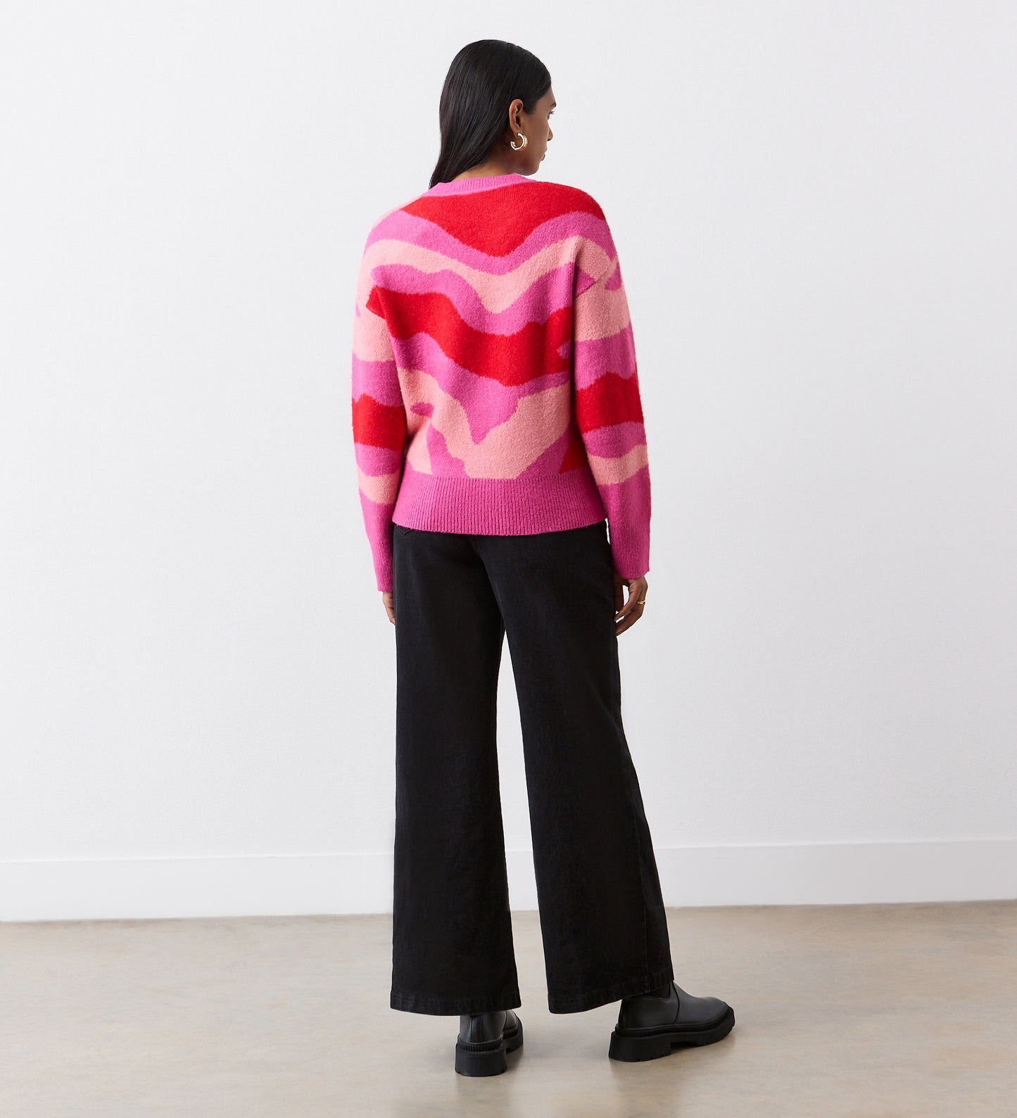 A person stands facing away, wearing the Minnie Pink Abstract Print Knitted Jumper with ribbed detailing, paired with black wide-leg pants and black platform boots. Their straight dark hair falls over their shoulders against a white wall.
