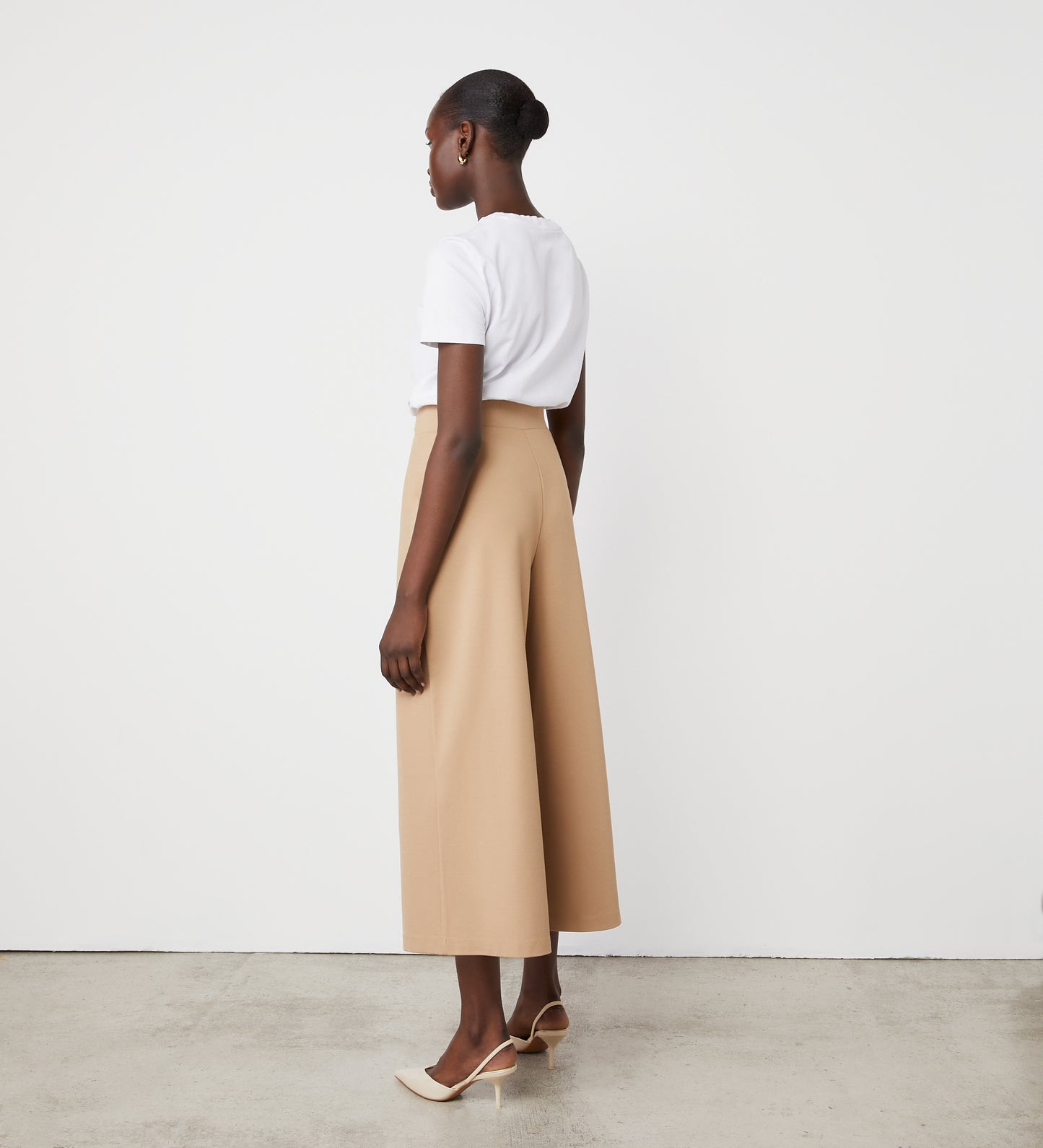 A woman stands sideways against a white wall, wearing a white short-sleeve top with Miribel Camel Cropped Ponte Jersey Trousers in a high-waisted, wide-leg fit, paired with beige heeled shoes. Her hair is styled in a low bun.