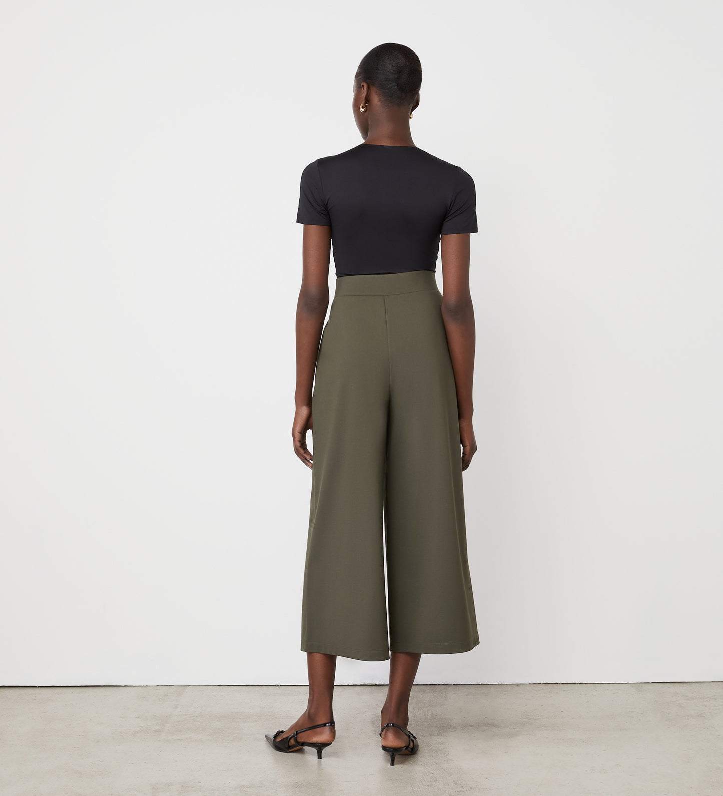 A person with short hair stands on polished concrete, wearing a fitted black short-sleeve top, black sandals, and high-waisted wide-leg Miribel Khaki Cropped Ponte Jersey Trousers while facing a white wall.