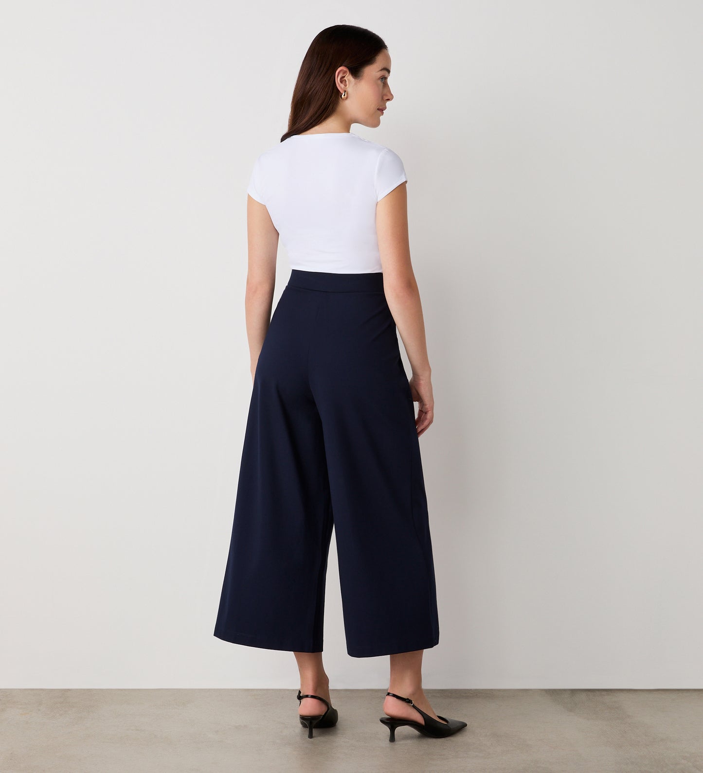 A woman faces a blank white wall, wearing a white fitted short-sleeve top and the Miribel Navy Cropped Ponte Jersey Trousers with black low-heeled shoes. She is shown from the back on a plain floor.