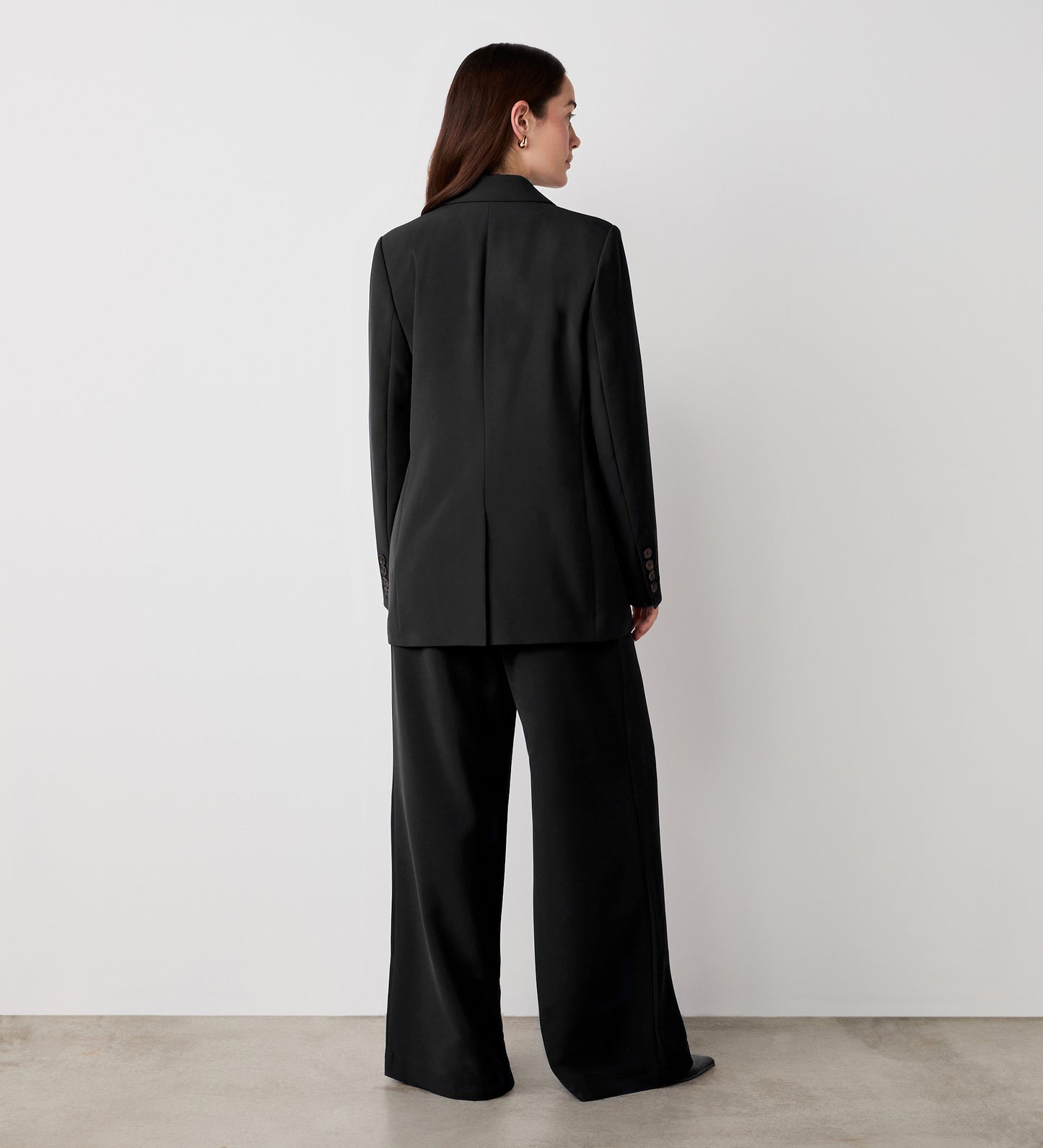 A person stands facing away from the camera, wearing the Myla Black Longline Blazer with wide-leg trousers, set against a plain white wall and concrete floor.
