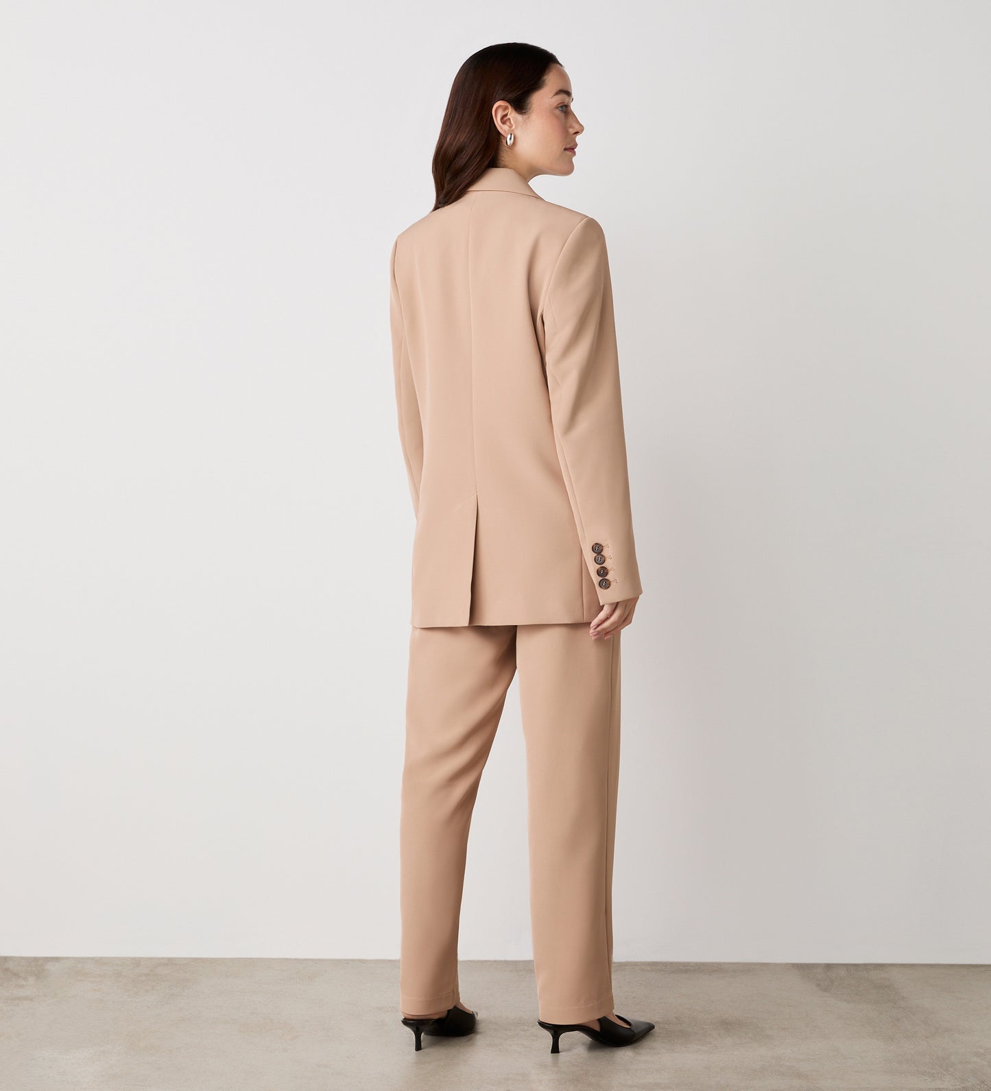 A woman with long brown hair wears the Myla Camel Longline Blazer, styled with relaxed fit trousers and black heeled shoes. She stands against a plain white wall on a concrete floor. The blazer features stylish functional pockets.