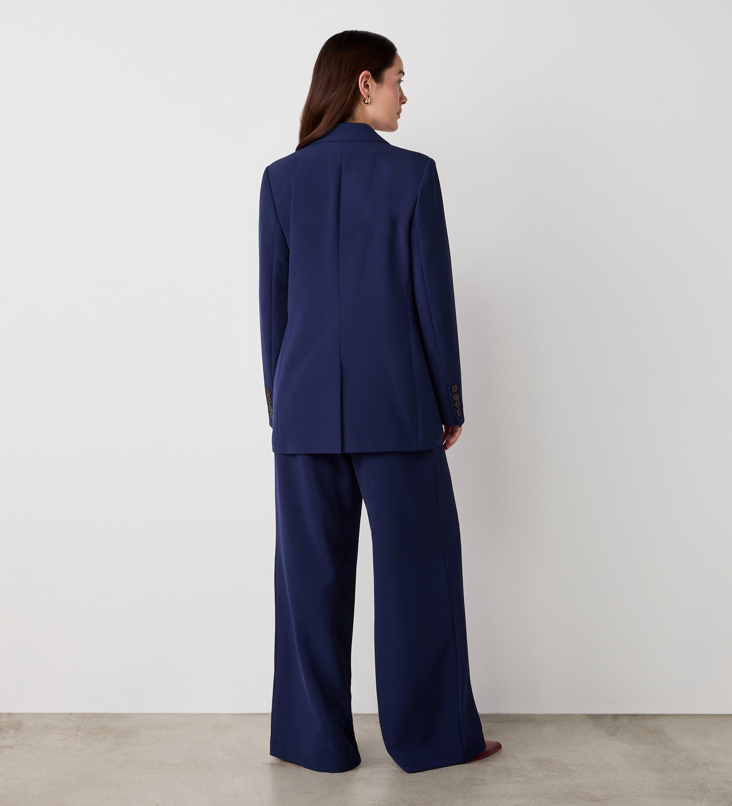 A woman faces away from the camera, wearing the Myla Navy Longline Blazer with a relaxed fit and wide-leg trousers, standing against a plain white wall and a light-colored floor.
