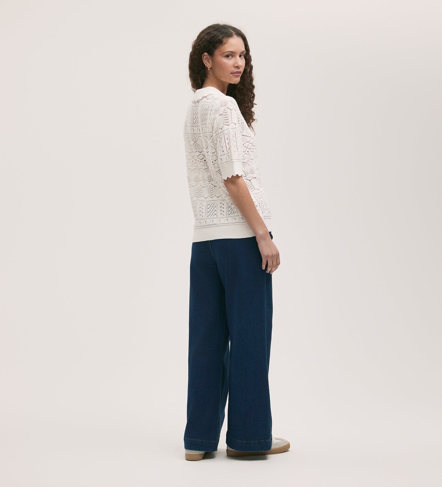 A woman with curly hair stands sideways, looking over her shoulder. She wears the Marlowe Cream Crochet Cardigan with wide-leg blue jeans and platform shoes, set against a plain light background.