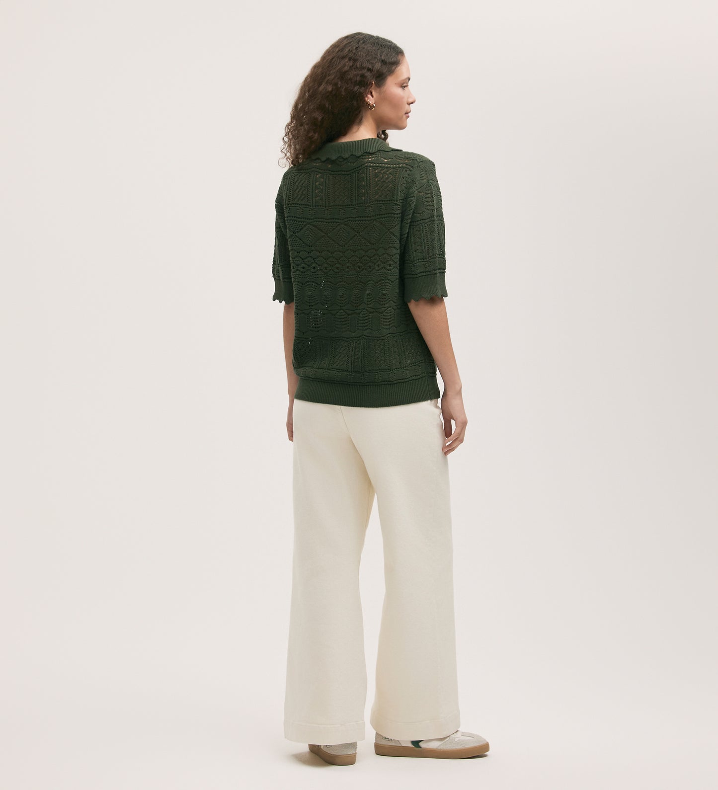 A woman stands facing away, wearing the Marlowe Khaki Crochet Cardigan with scallop detailing, wide-leg white pants, and beige platform shoes against a plain light background.