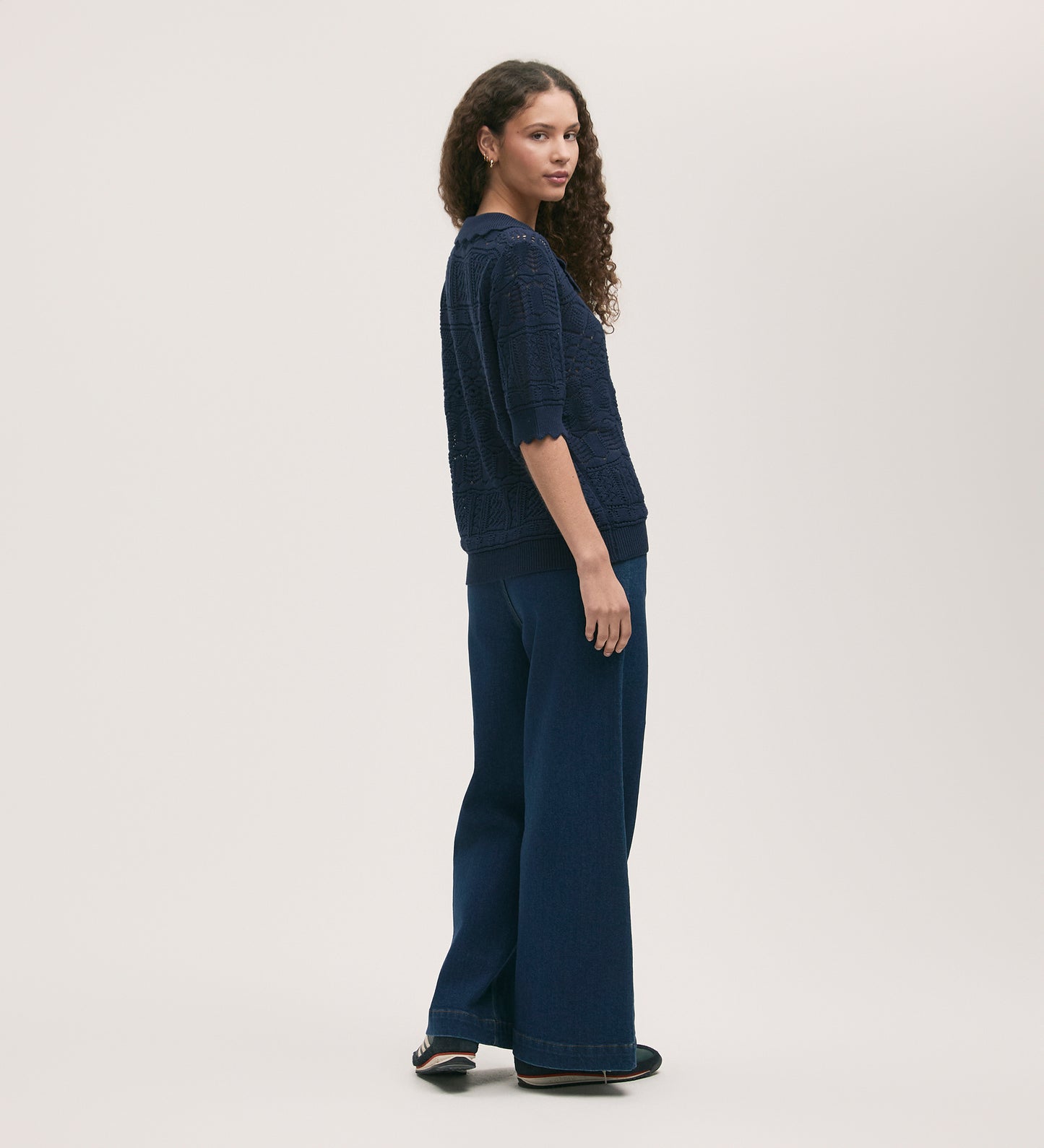 A woman with long curly hair wears the Marlowe Navy Crochet Cardigan, dark blue wide-leg pants, and black shoes in a neutral studio setting, looking over her shoulder.