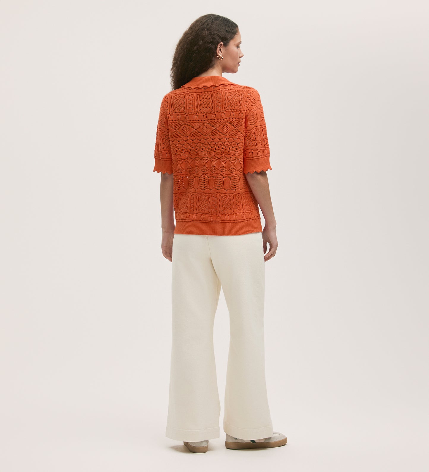 A person with long curly hair, wearing the Marlowe Orange Crochet Cardigan and high-waisted, wide-leg white pants, stands facing away against a plain light background.