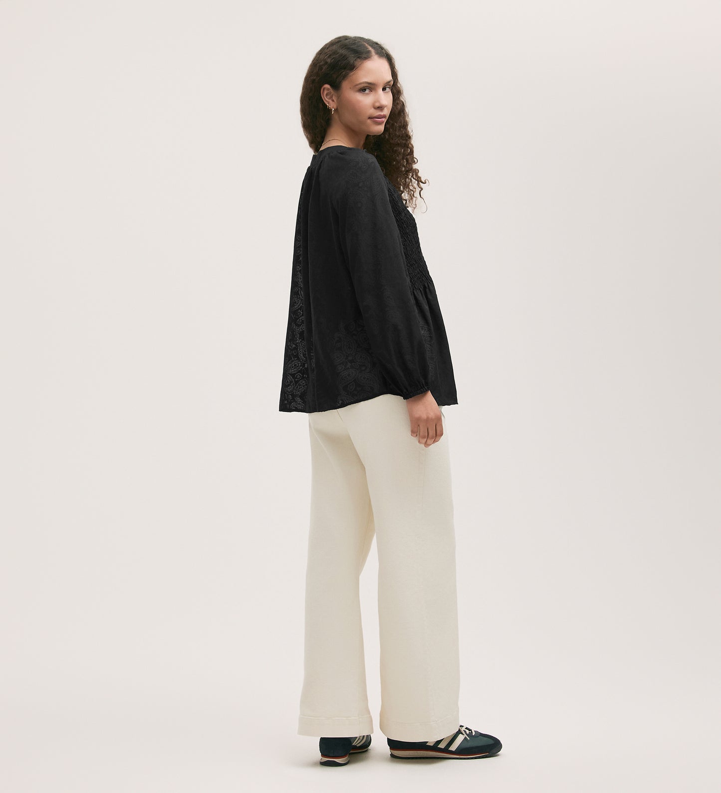 A woman with curly hair stands in profile wearing a loose Martha Black Chiffon Blouse with long sleeves and wide-leg cream pants, paired with black shoes, against a plain light background.