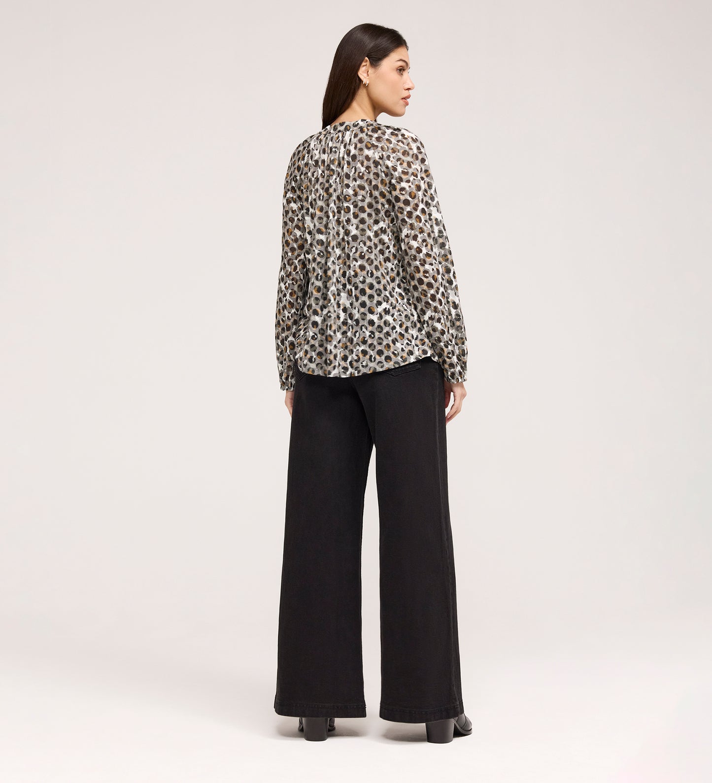 A woman faces away, wearing the Martha Khaki Animal Jersey Blouse with long sleeves and leopard print, paired with wide-leg black pants and platform shoes, set against a plain light background.