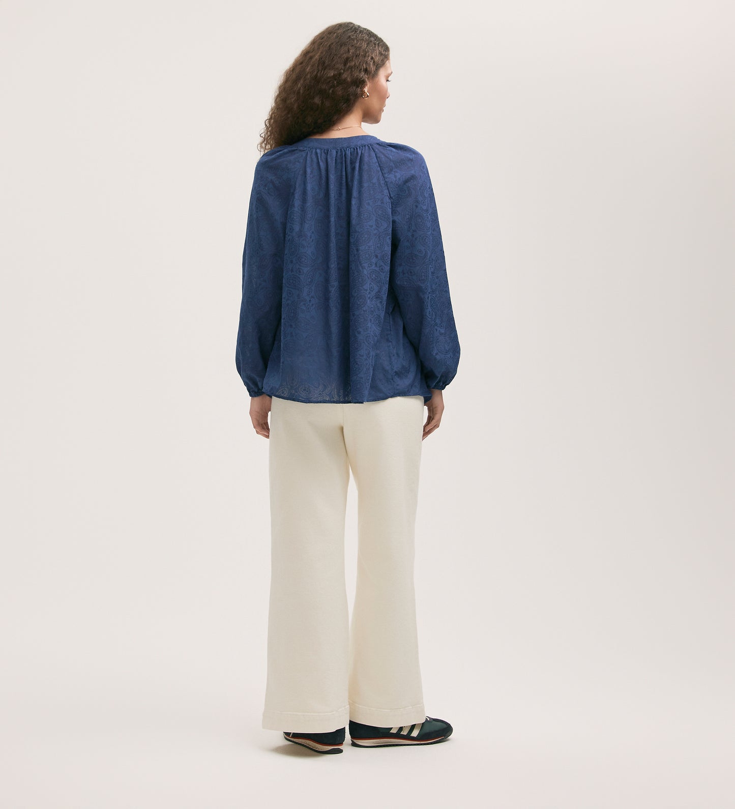 A person with long, curly hair faces away, wearing a loose Martha Navy Chiffon Blouse with a shirred bodice, wide-leg white pants, and black sneakers with white stripes against a plain light background.