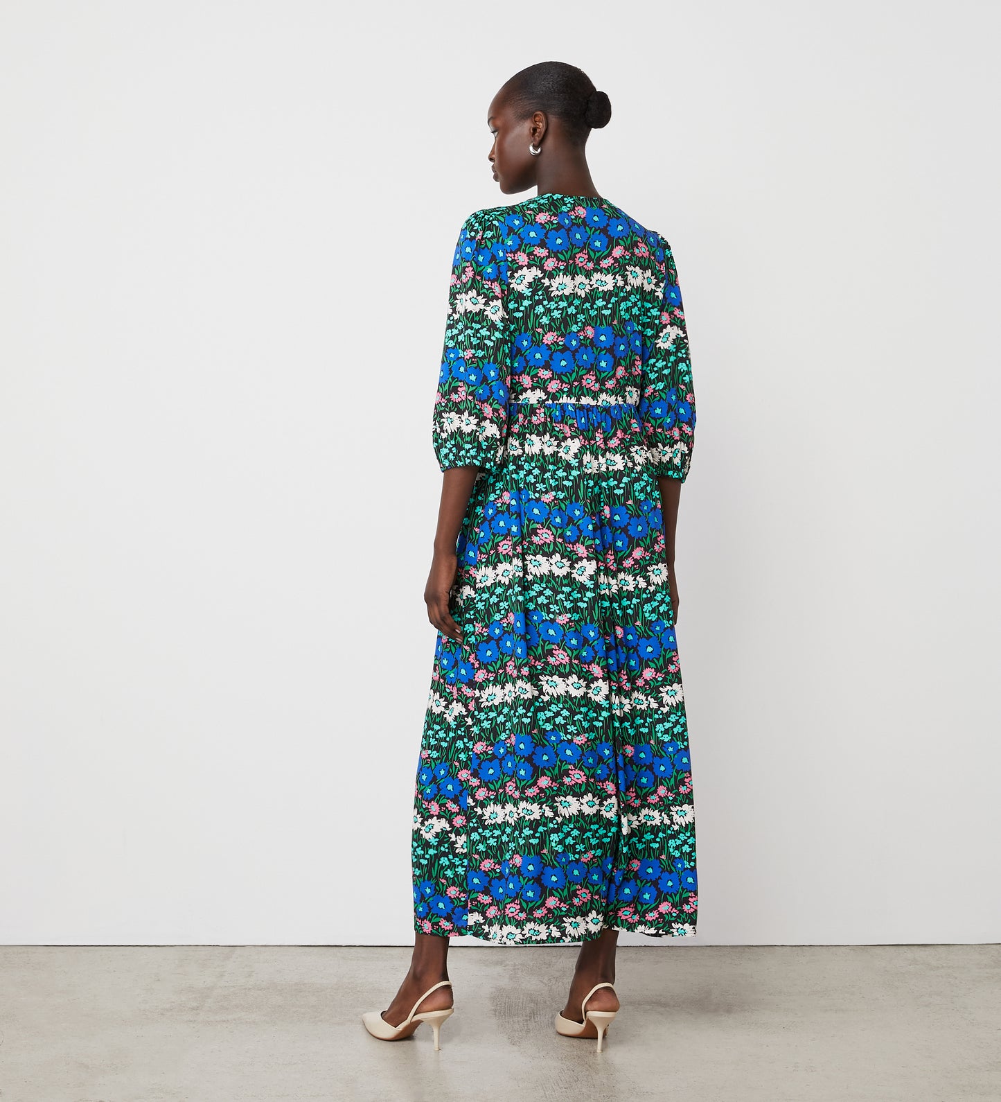 A woman faces away in the Naomi Blue Floral Stripe Crepe Midi Dress with three-quarter sleeves and white heels, set against a plain white wall and concrete floor.