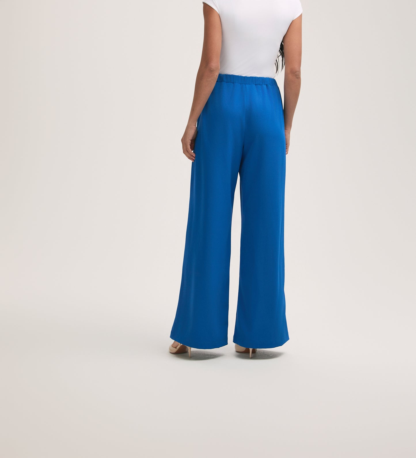 A woman stands facing away, wearing a white short-sleeve top and Nat Cobalt Blue Crepe Wide Leg Trousers with an elasticated waist. She is standing on a light, plain background.