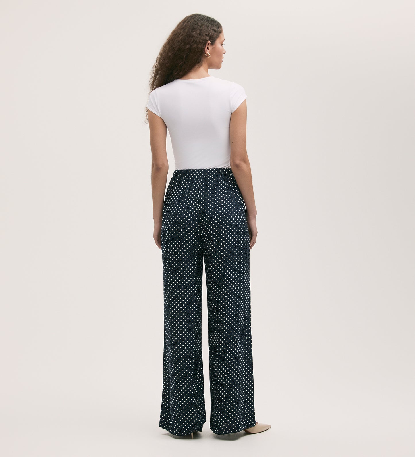 A woman with long curly hair faces away, wearing a white fitted tee and Nat Navy Polka Dot Wide Leg Trousers, made from crepe fabric with a high waist and white polka dots. The background is plain and light.