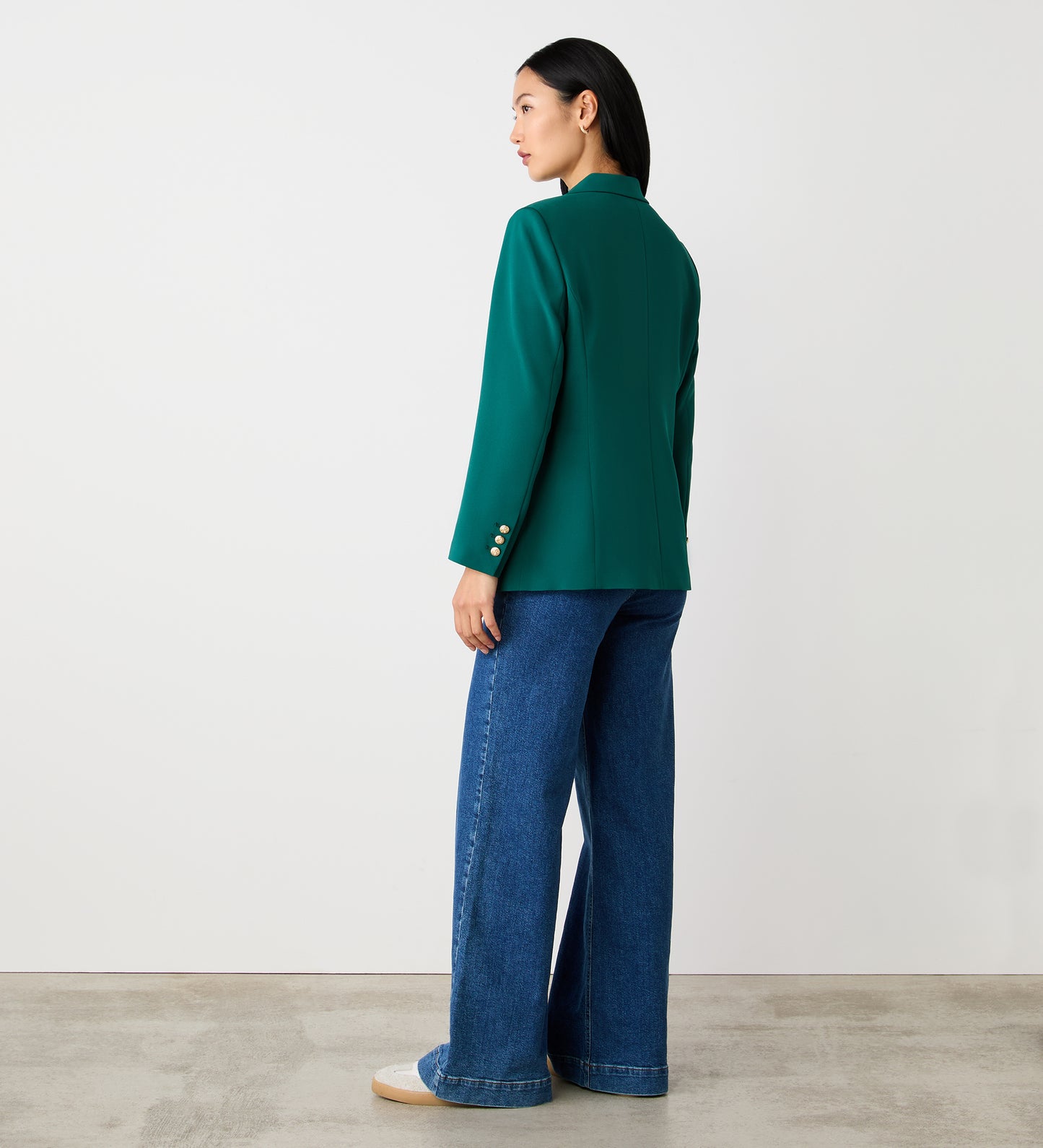 A woman with straight black hair stands facing away, wearing a tailored Orla Forest Green Blazer with gold buttons and wide-leg blue jeans, set against a plain white wall and concrete floor.