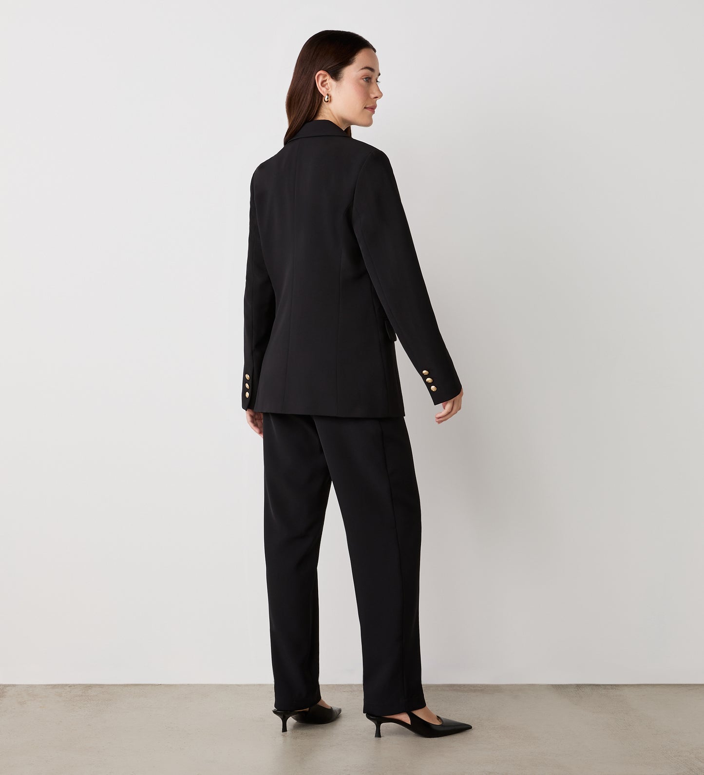 A woman with straight brown hair faces away, wearing the Orla Black Tailored Blazer with gold button details and matching trousers, paired with black heels, against a plain white wall and concrete floor.