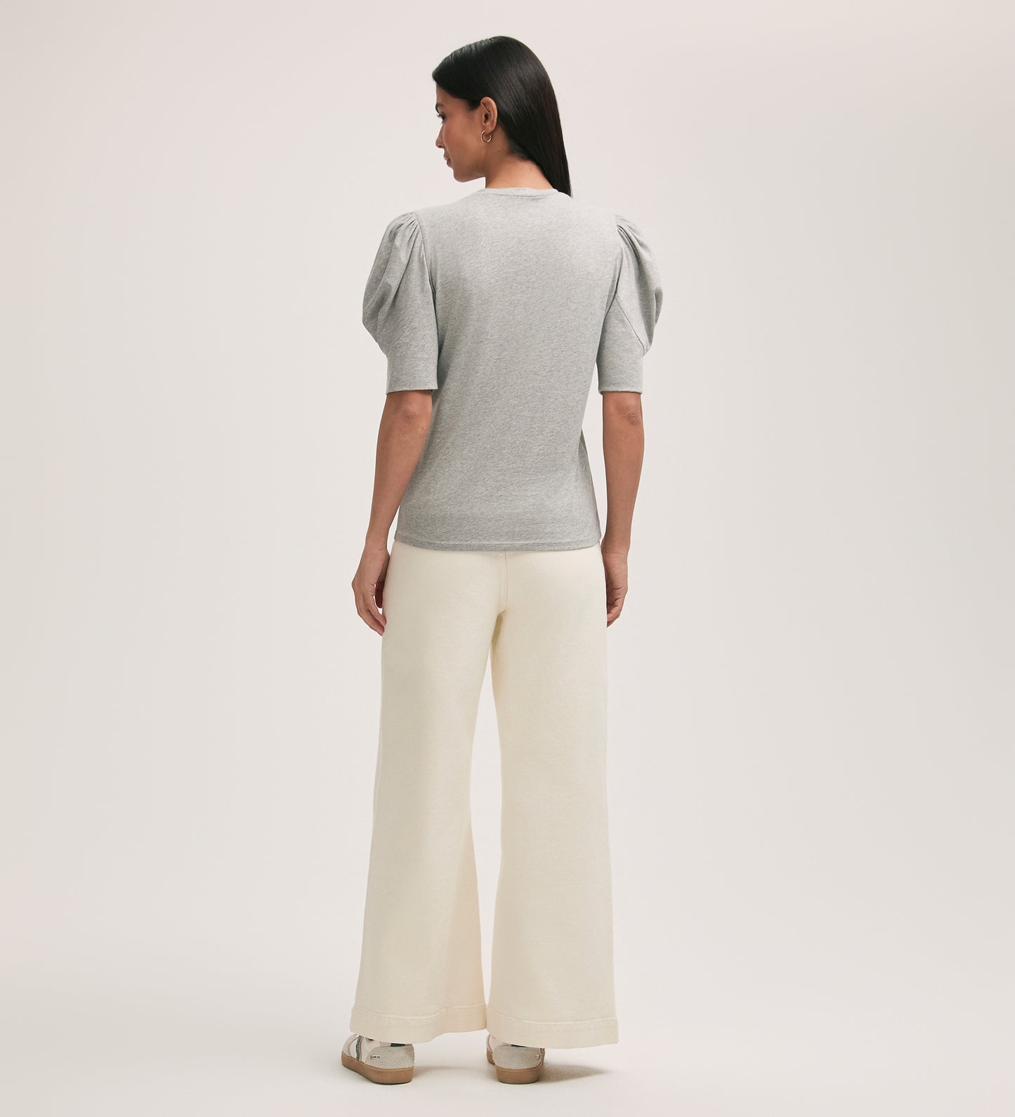 A woman with straight dark hair faces away, wearing the Oriel Grey Jersey T-Shirt with puffed short sleeves and wide-leg cream pants, styled with light-colored platform sandals against a plain background.