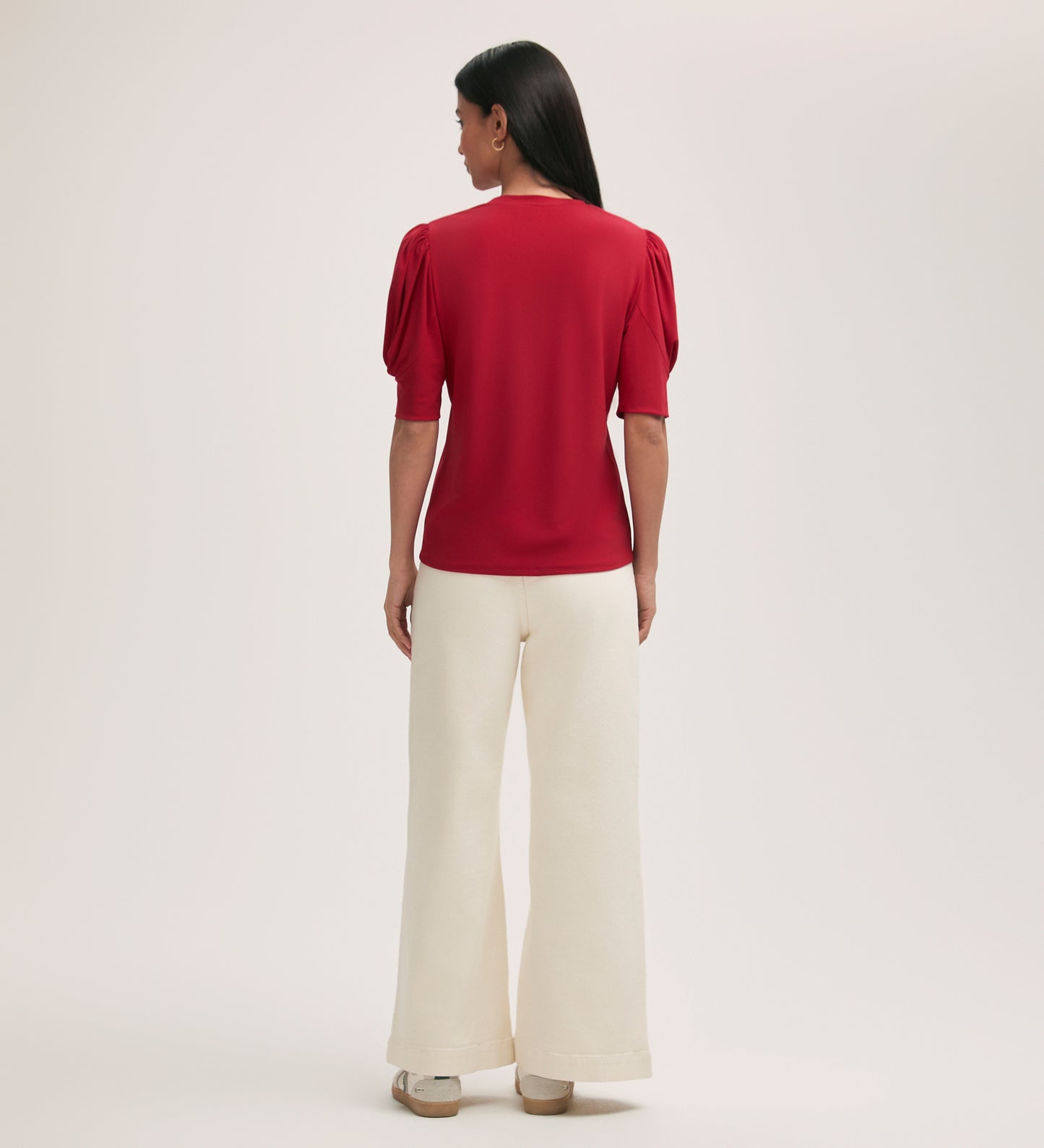 A woman faces away wearing the Oriel Red Jersey T-Shirt with puffed shoulders, styled with wide-leg, high-waisted white pants and beige shoes. The setting has a plain, light-colored background.